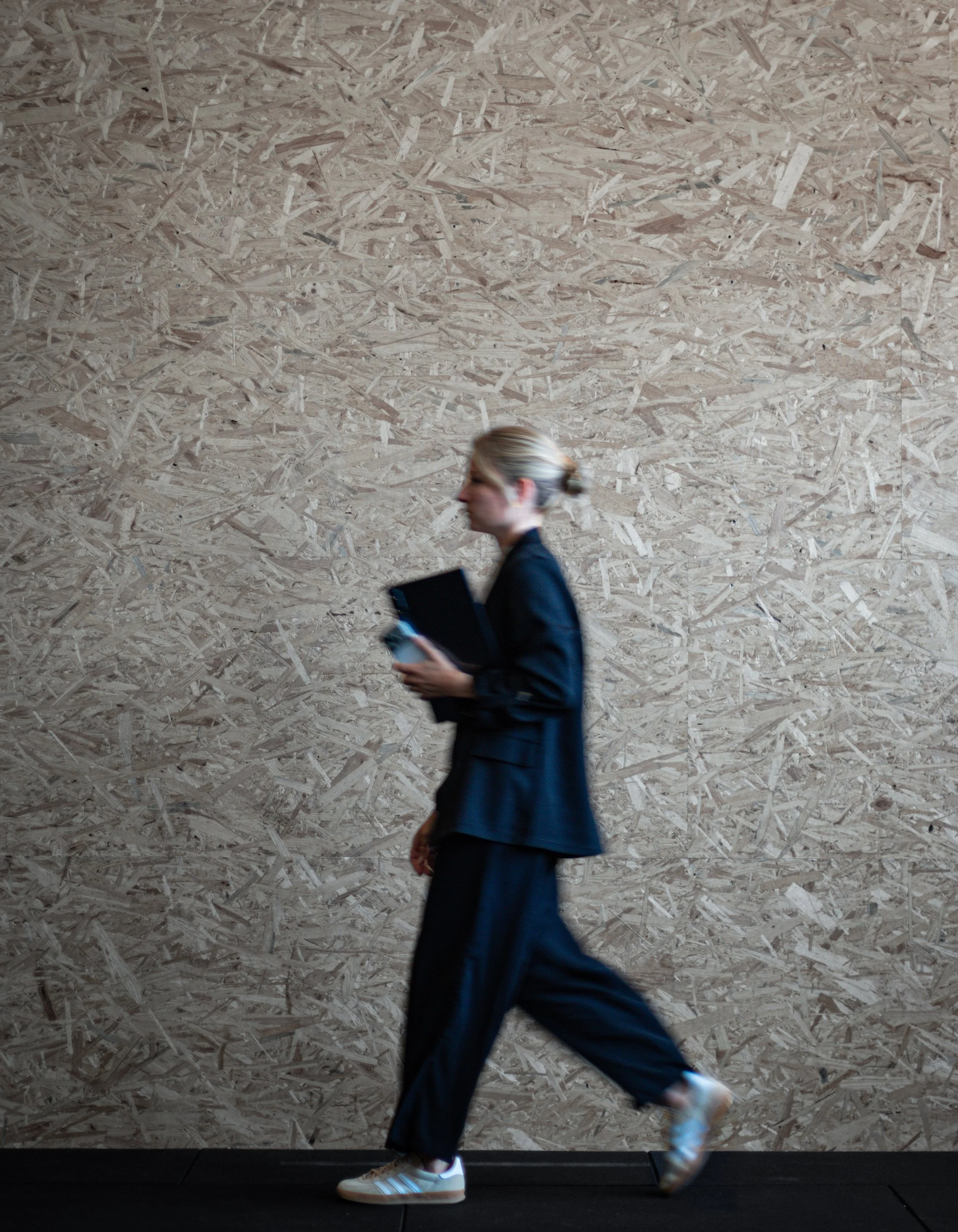 Eine Frau läuft an einer Holzwand mit Schnittholzoptik vorbei, trägt eine schwarze Jacke, schwarze Hosen, weiße Turnschuhe und hält ein Notebook sowie ein Smartphone in den Händen.