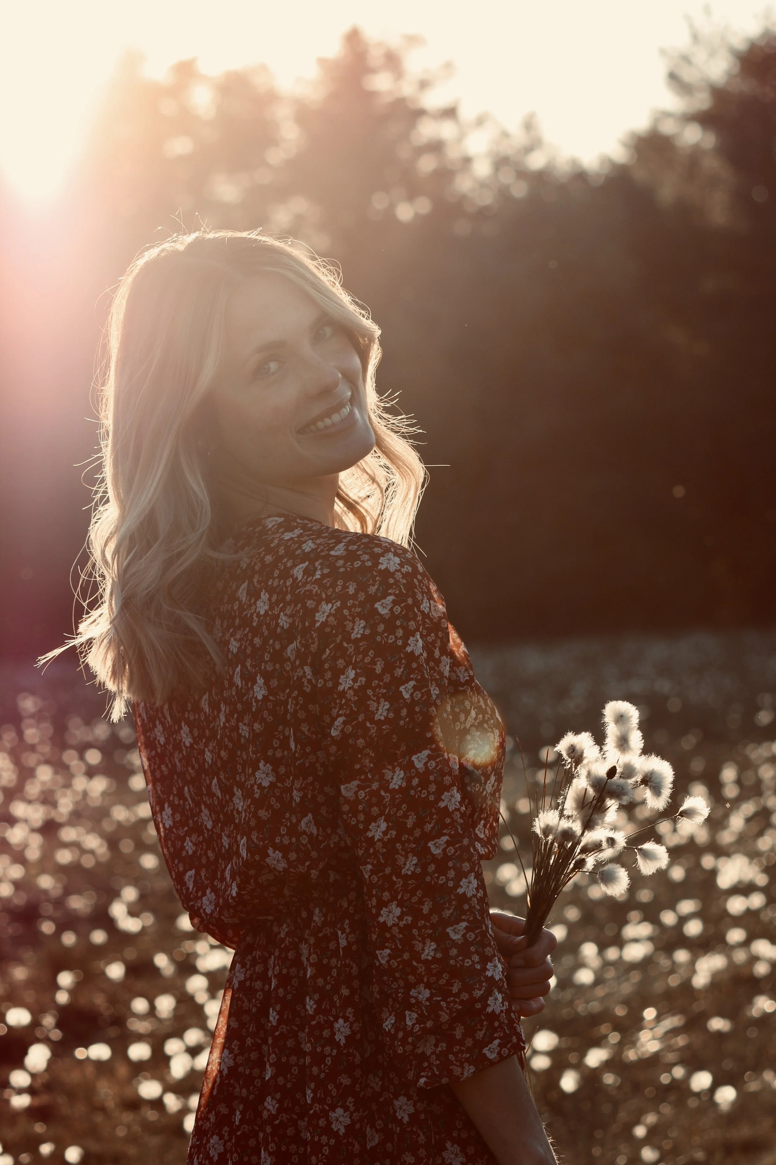 Eine junge Frau mit blonden Haaren trägt ein rotes, gemustertes Kleid und hält einen Bund von weißen Pusteblumen in der Hand. Sie steht im Sonnenlicht, im Hintergrund sind Wasser und Bäume sichtbar.