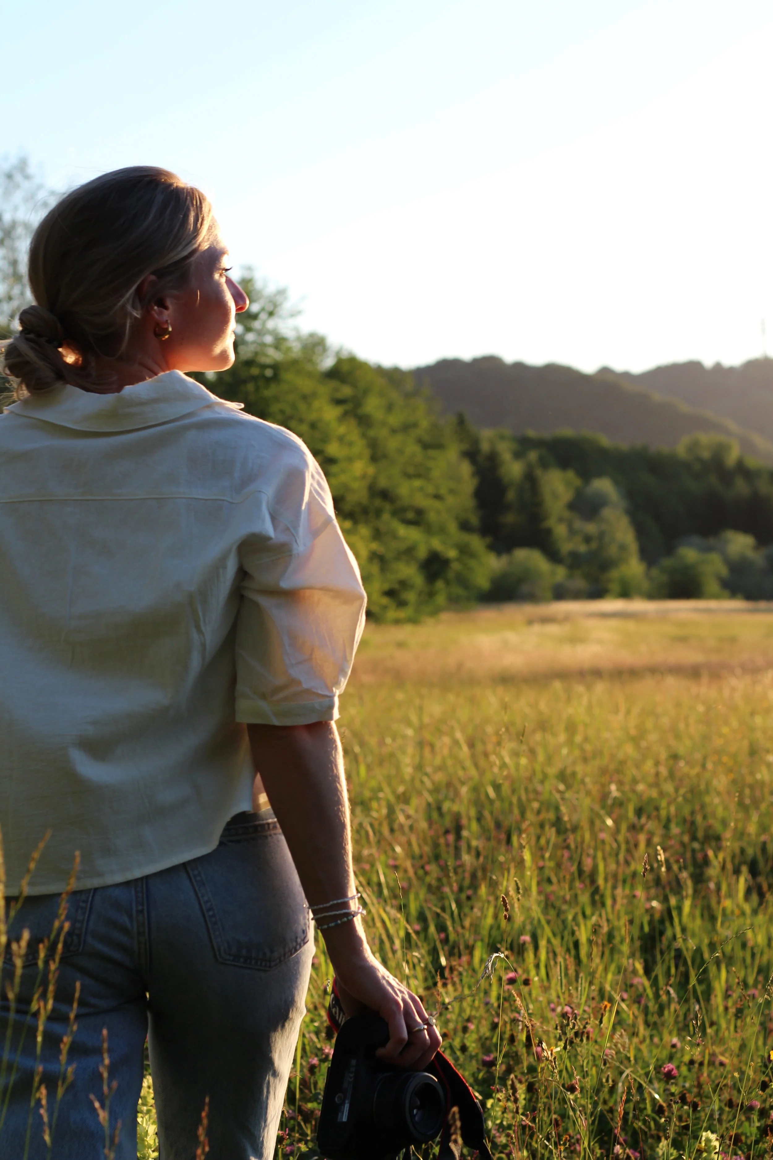 Frau steht in einem Feld bei Sonnenuntergang, blickt nach rechts und hält eine Kamera in der Hand.
