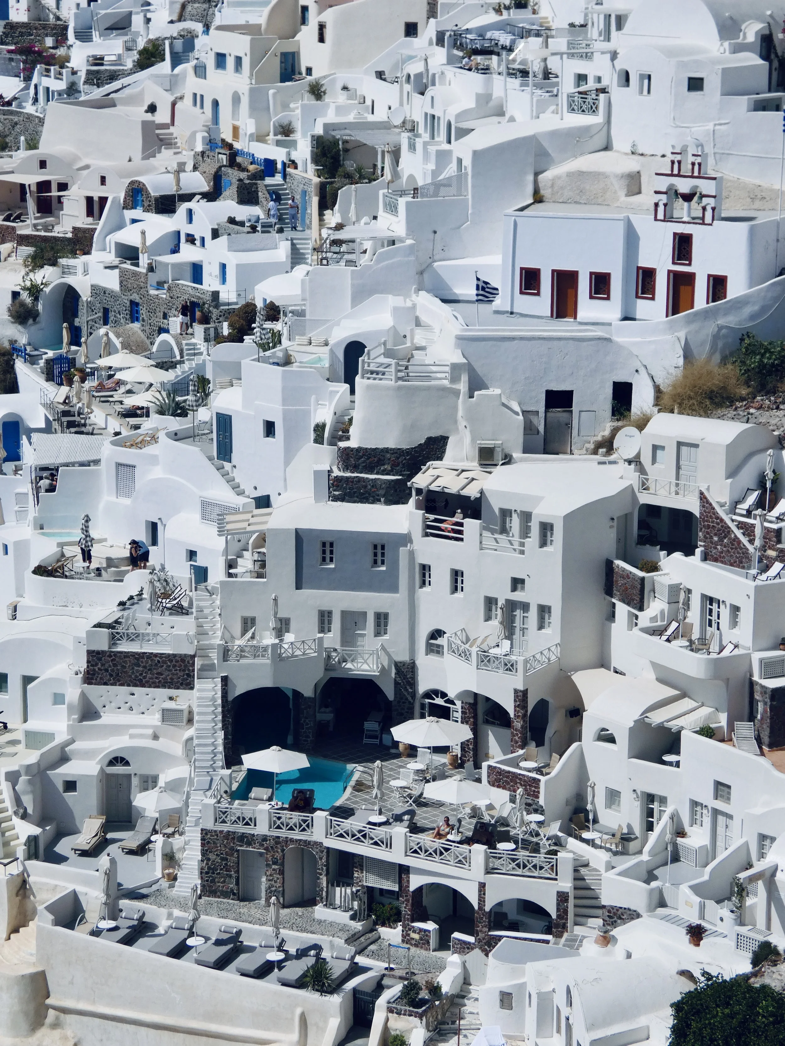 Blick auf weiße, terrassenförmig angeordnete Häuser im Stil der Kykladen auf Santorini, Griechenland, mit Balkonen, Sonnenschirmen, Pool und Urlaubern.