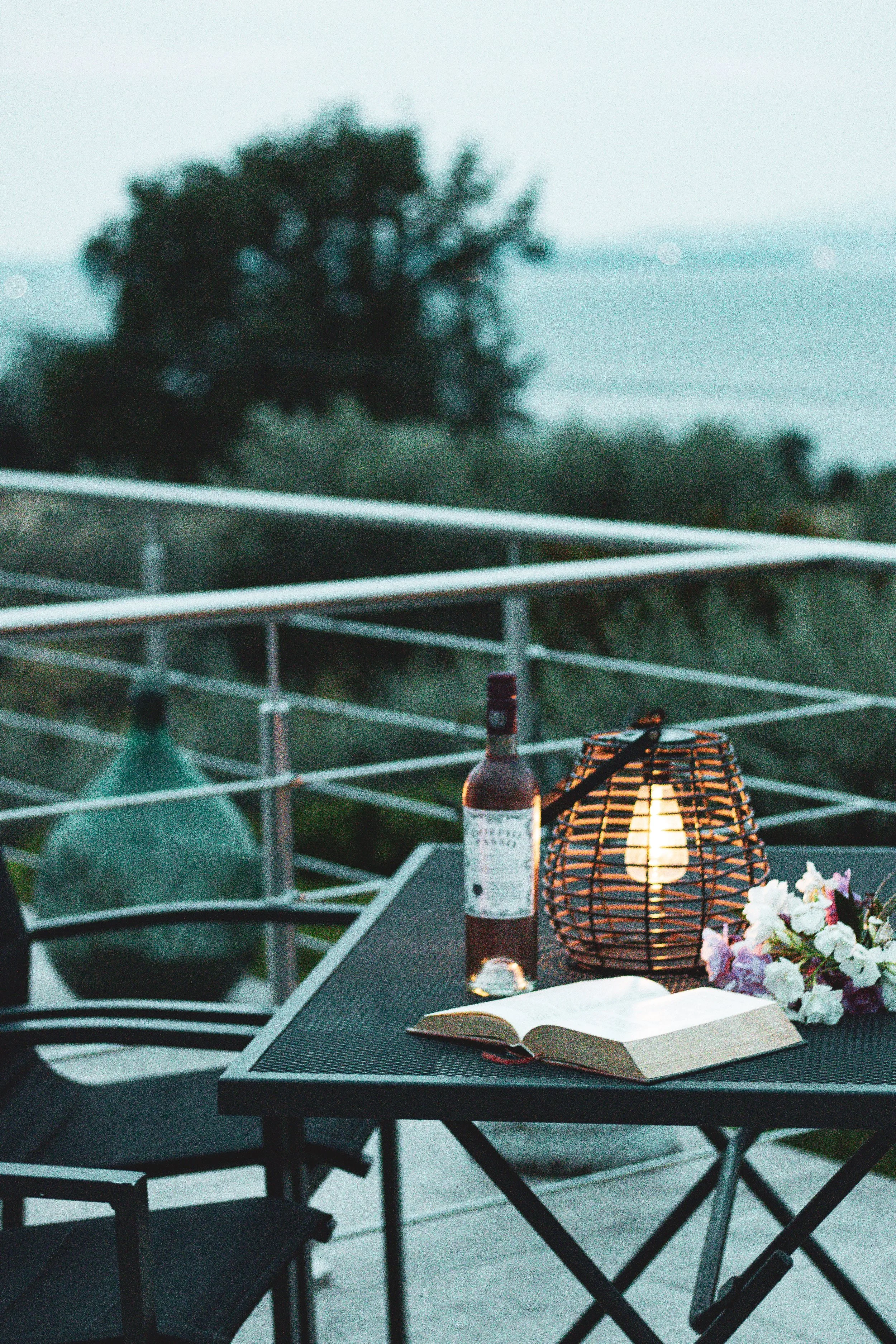 Auf einem Balkon steht ein Tisch mit einem offenem Buch, einer Flasche Wein, einem Kerzenleuchter mit brennender Kerze, einer Laterne mit glühender Glühbirne, einem kleinen Blumenstrauß und einer weiteren Kerze. Im Hintergrund sind Bäume und das Meer sichtbar, es ist dämmerig.