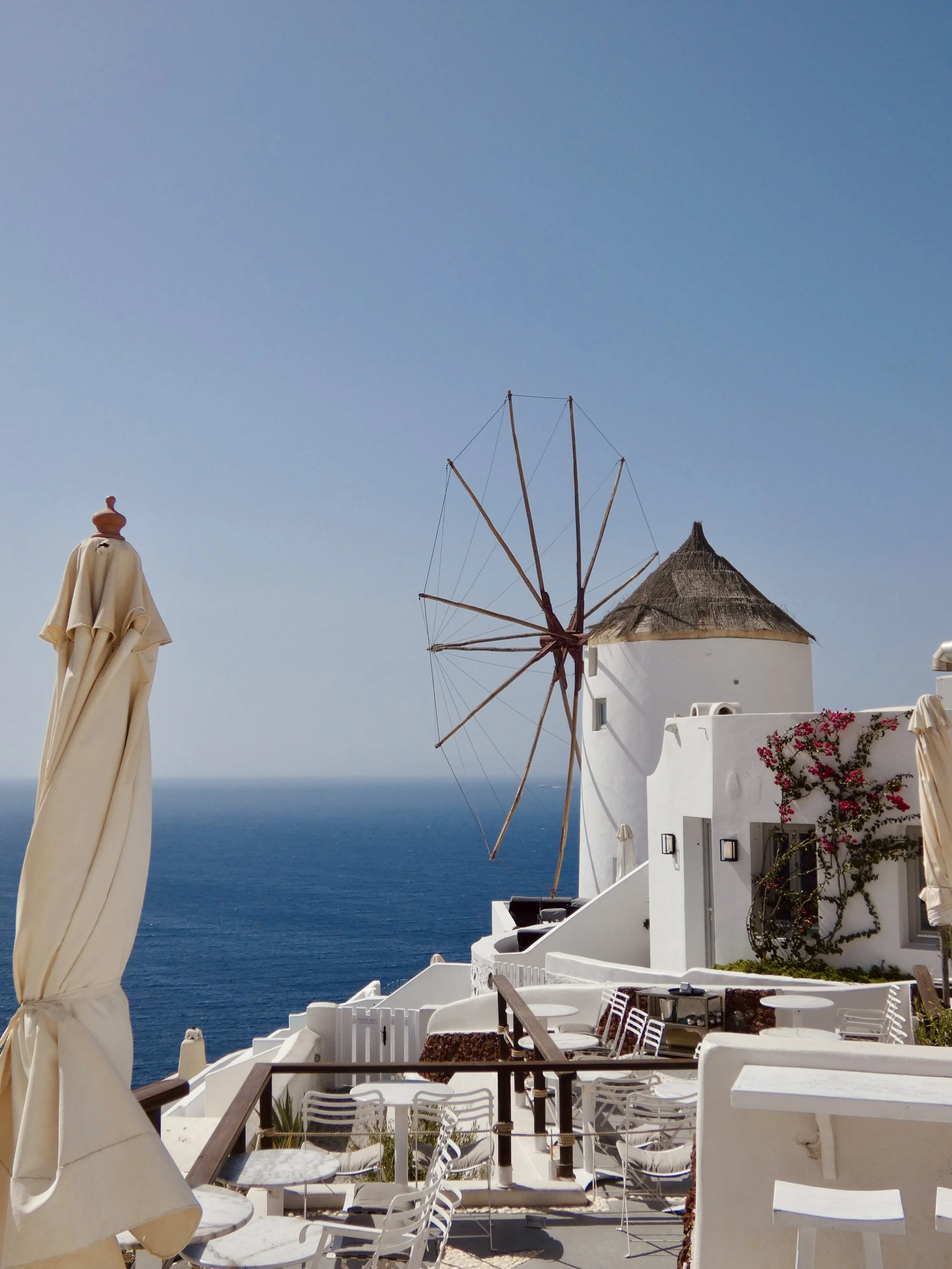 Blick auf eine Terrasse mit weißen Möbeln, Sonnenschirm, einer Windmühle und Blick auf das Meer in Santorini, Griechenland.