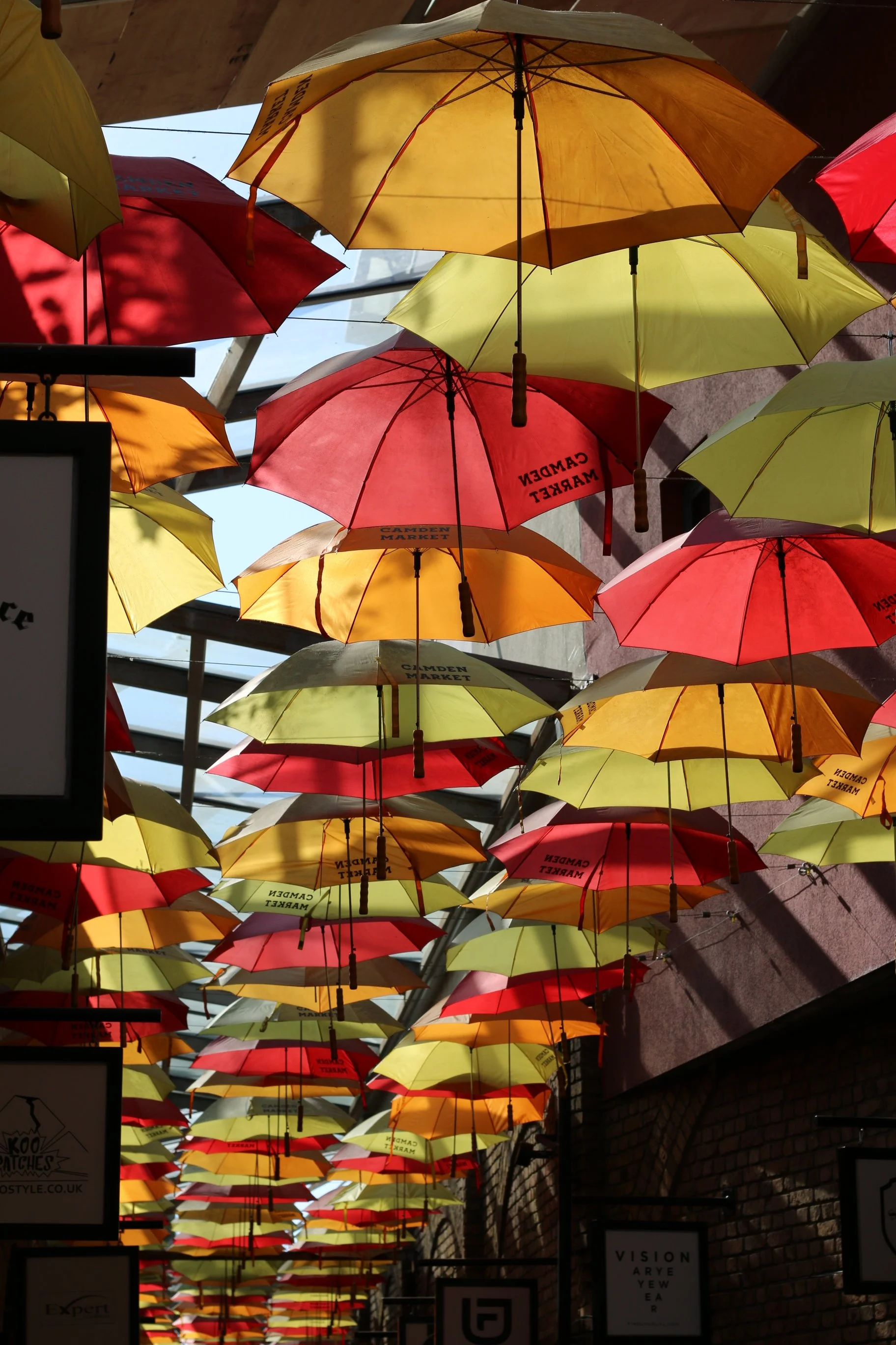 Bunte Regenschirme hängen an der Decke eines Unterstands, Schatten fällt auf die Schirme und die Innenwand aus Ziegelstein.