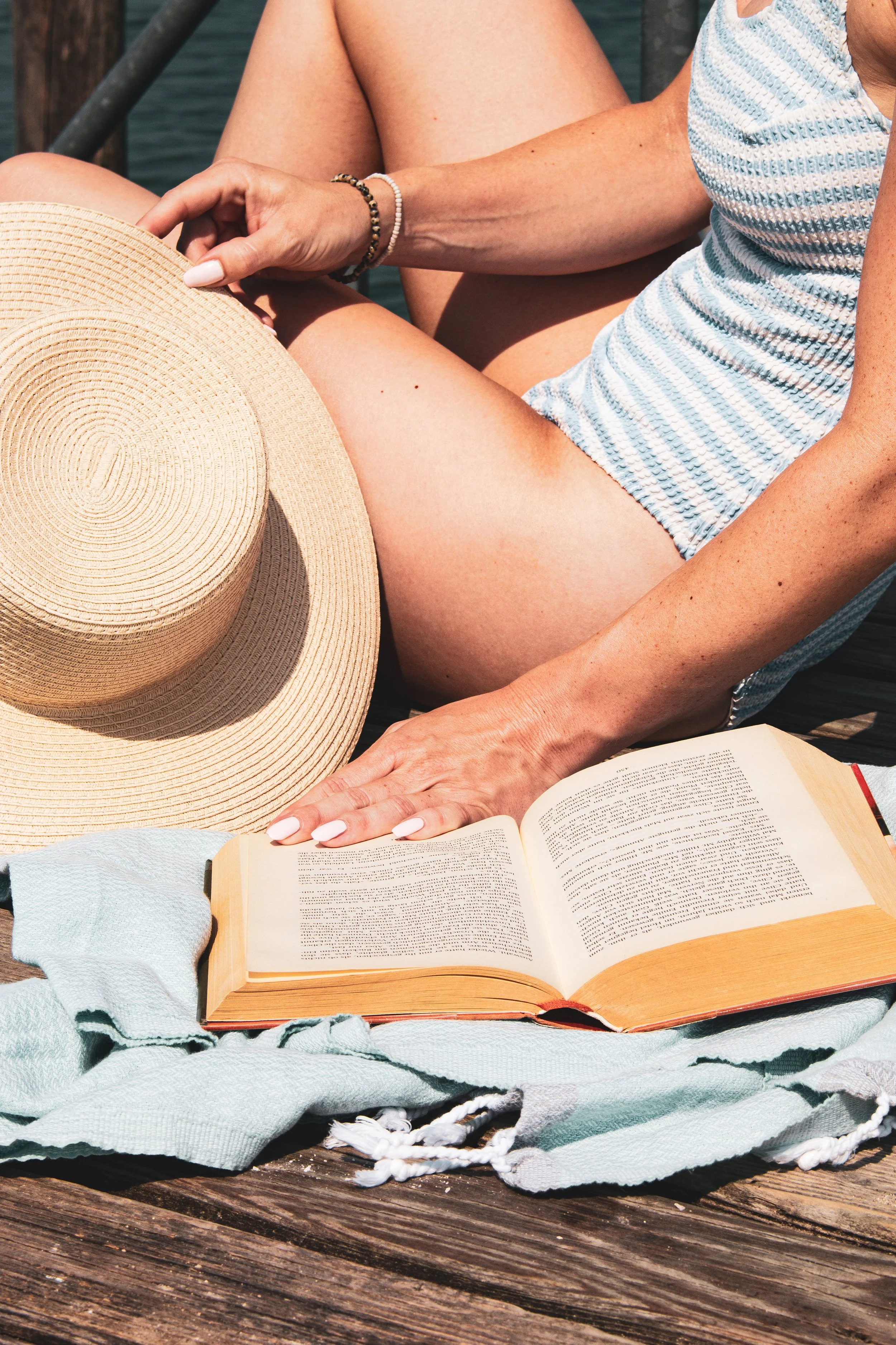 Nahaufnahme einer Person, die ein Buch liest, während sie im Freien auf einem Holzsteg sitzt, mit Sonnenhut, Strandtuch und Wasser im Hintergrund.