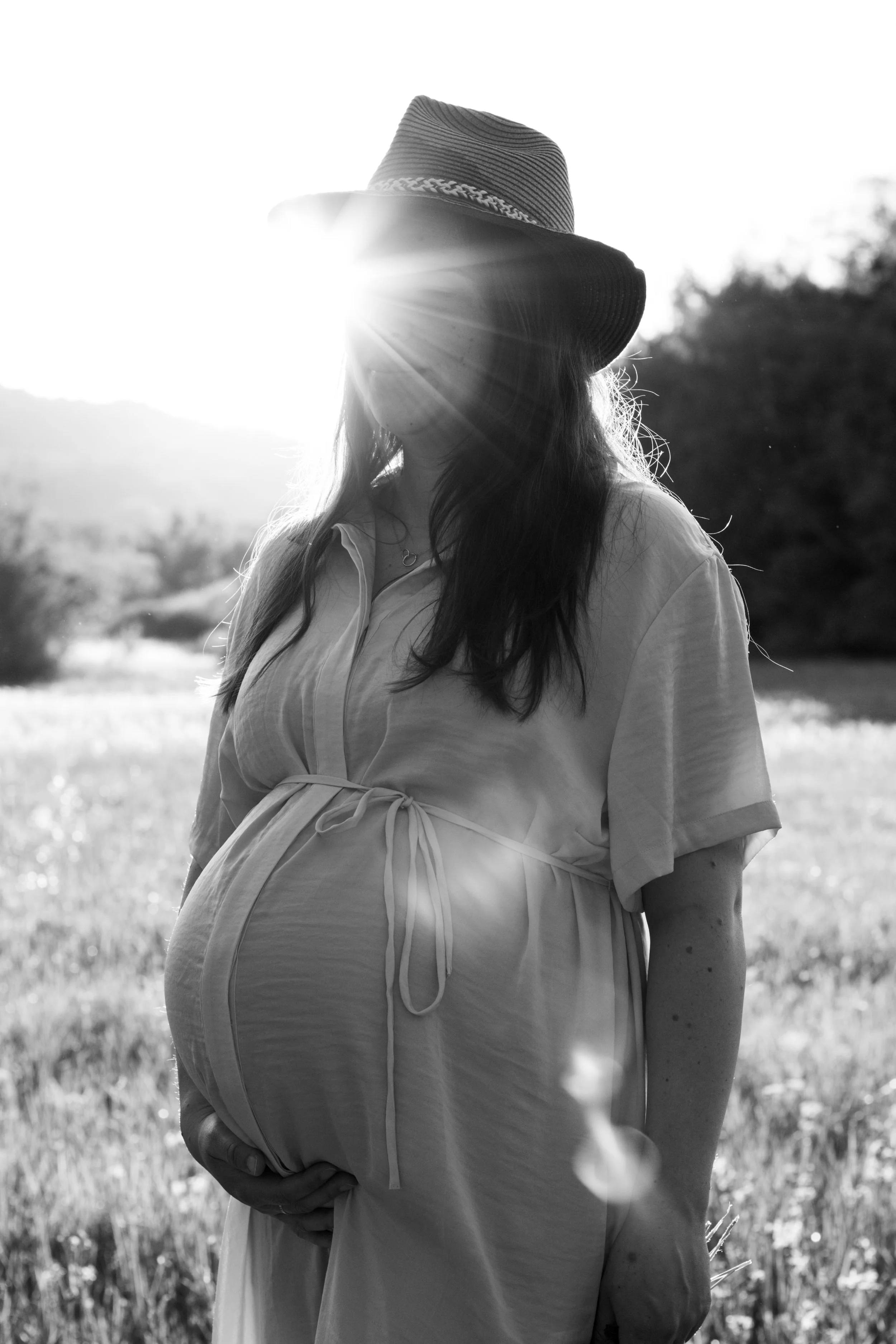 Schwarz-Weiß-Foto einer schwangeren Frau, die im Freien bei Sonnenuntergang steht, trägt einen Hut und ein lockeres Kleid, ihre Hand ruht auf ihrem Bauch.