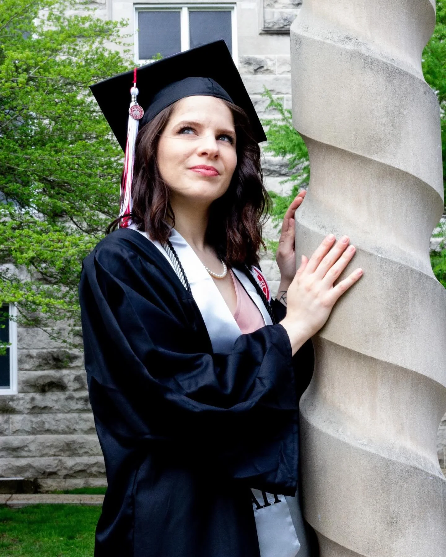 A woman in graduation attire with a black cap and gown, holding a spiral stone sculpture outdoors with green trees and a stone building in the background.