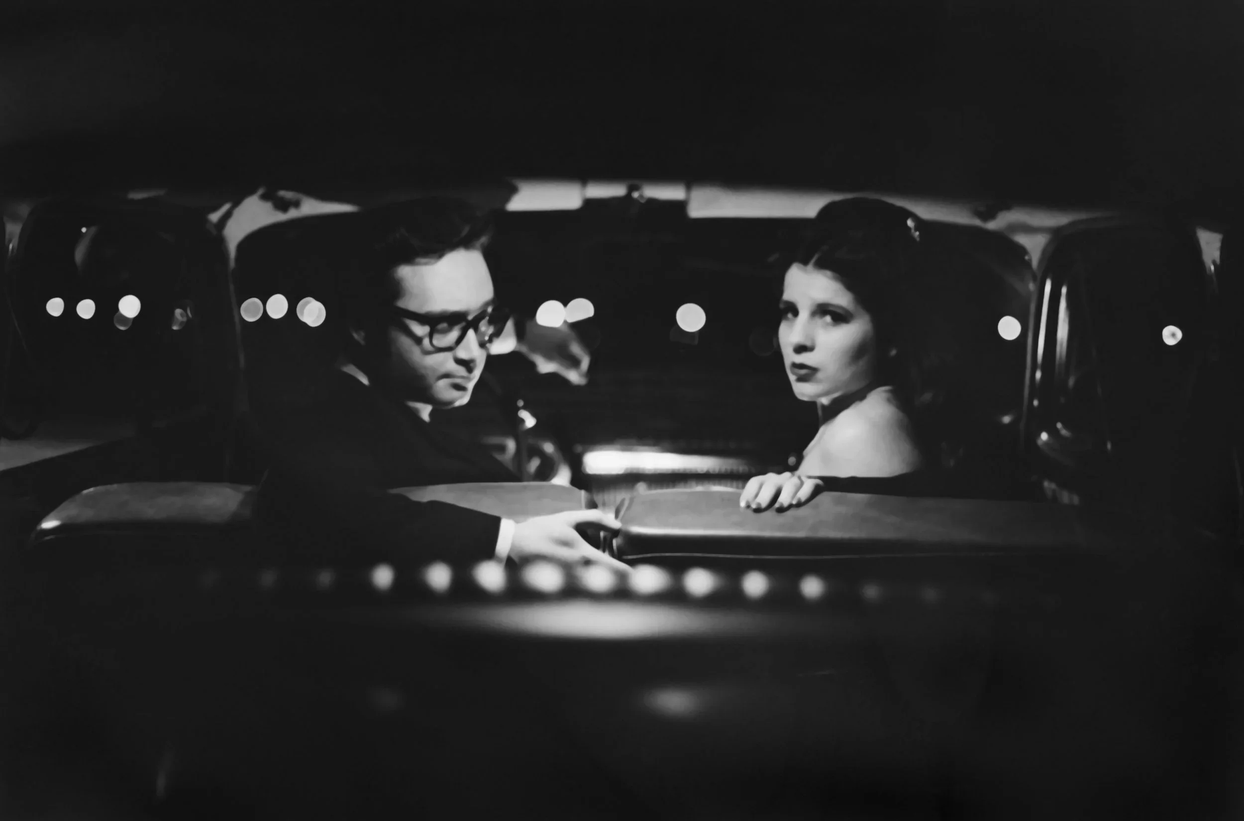 A black-and-white photo of a man and woman sitting in the backseat of a car at night, looking towards the camera.