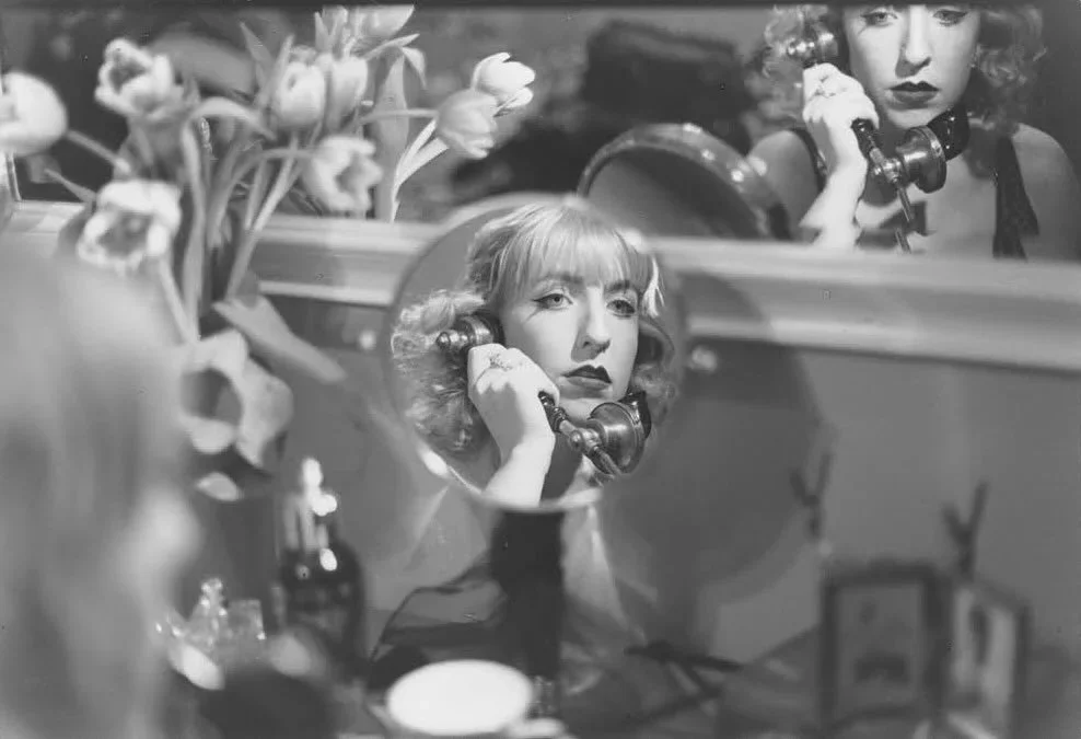 A woman with curly hair and dark lipstick talking on an old-fashioned telephone, her reflection visible in a mirror.