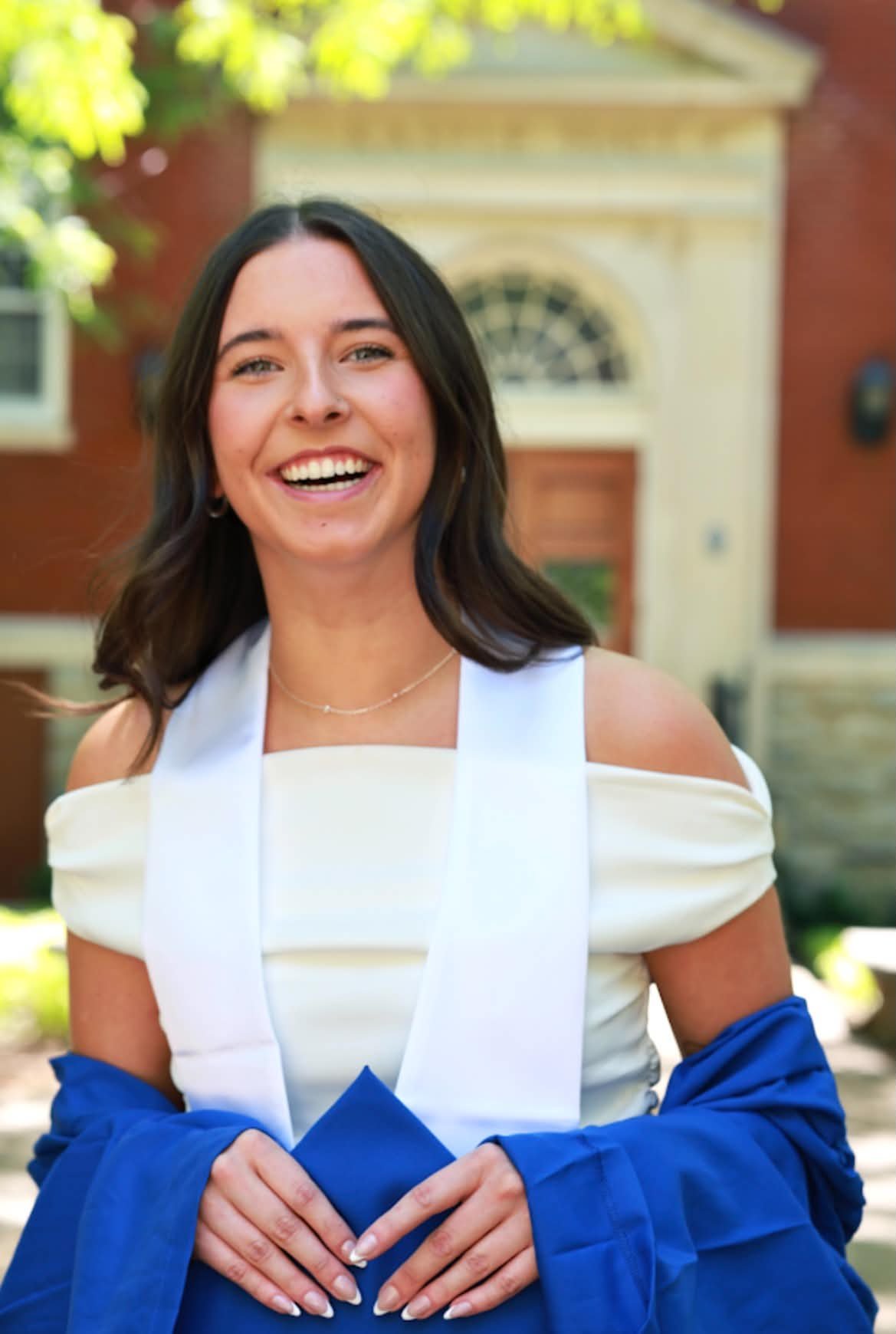 Young woman smiling outdoors, wearing a white dress and holding a blue jacket, with a building and greenery in the background.
