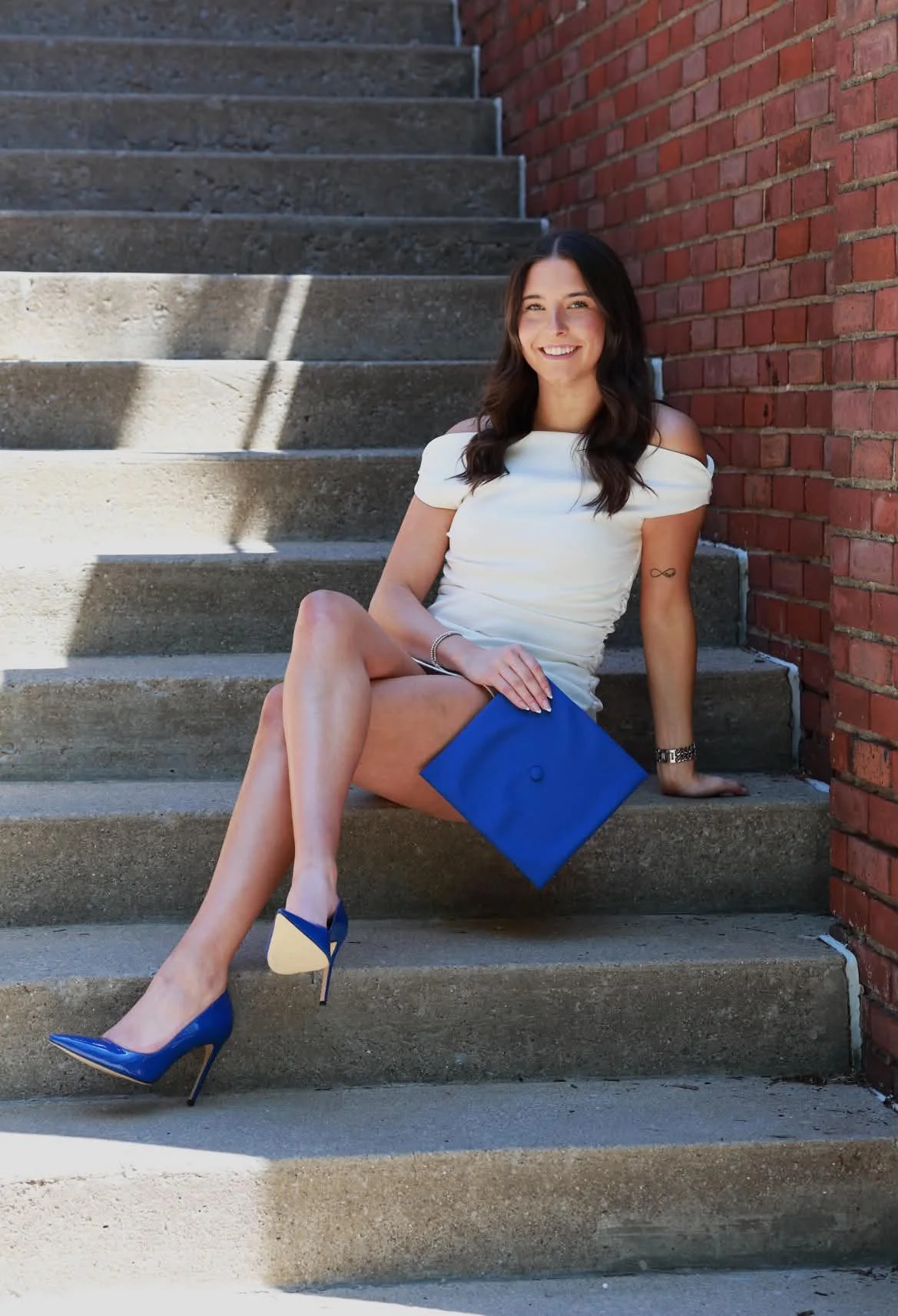A young woman with dark hair sitting on concrete stairs, leaning against a red brick wall, smiling, wearing blue high heels, a white off-shoulder top, and a blue skirt, holding a matching blue clutch.