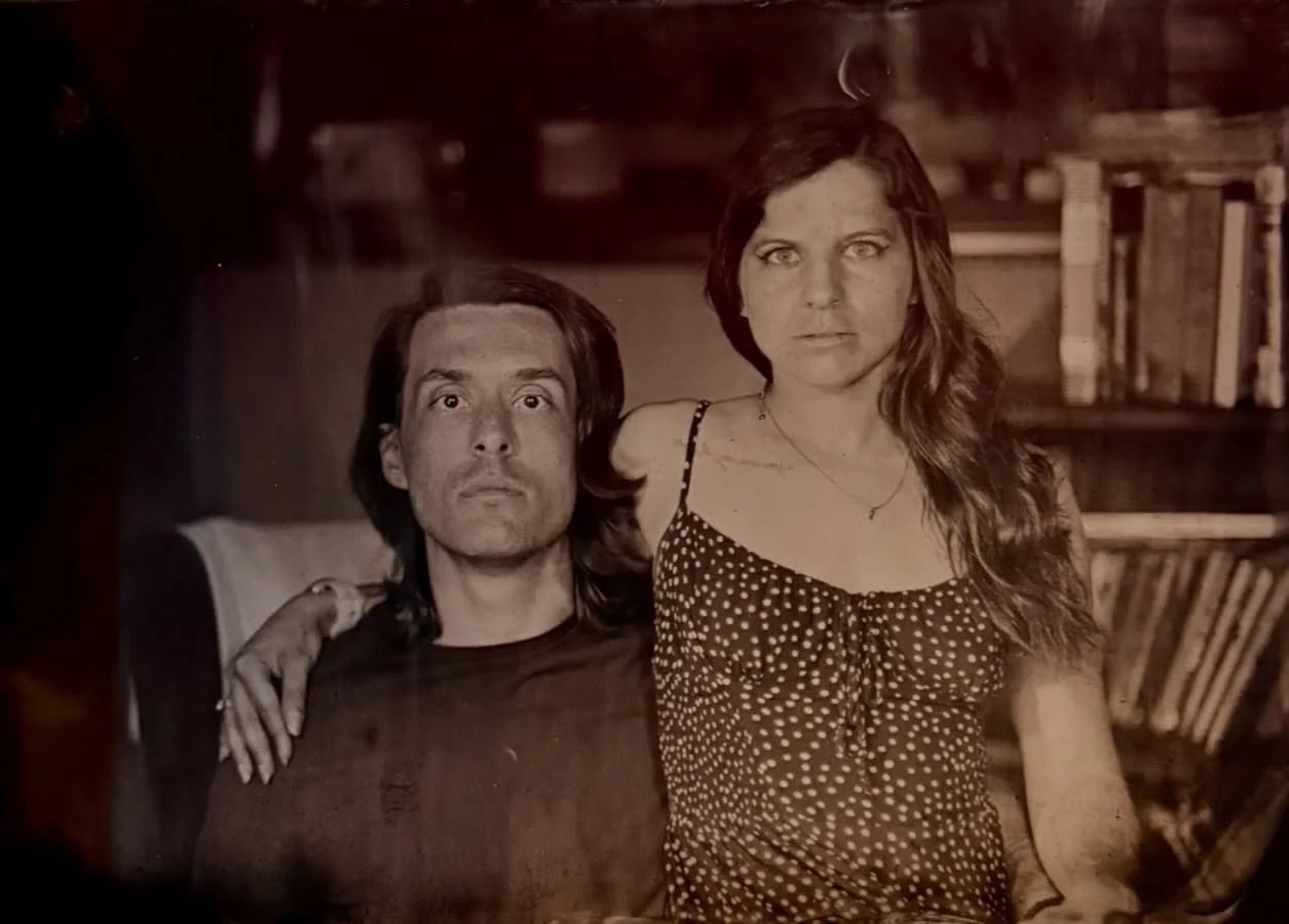 A black and white photo of a man and a woman sitting closely together, with the woman's arm around the man's shoulder. Both are looking directly at the camera with serious expressions. The woman has long, wavy hair and is wearing a polka-dot dress, while the man has straight hair and is wearing a dark shirt. In the background, there are shelves with books.