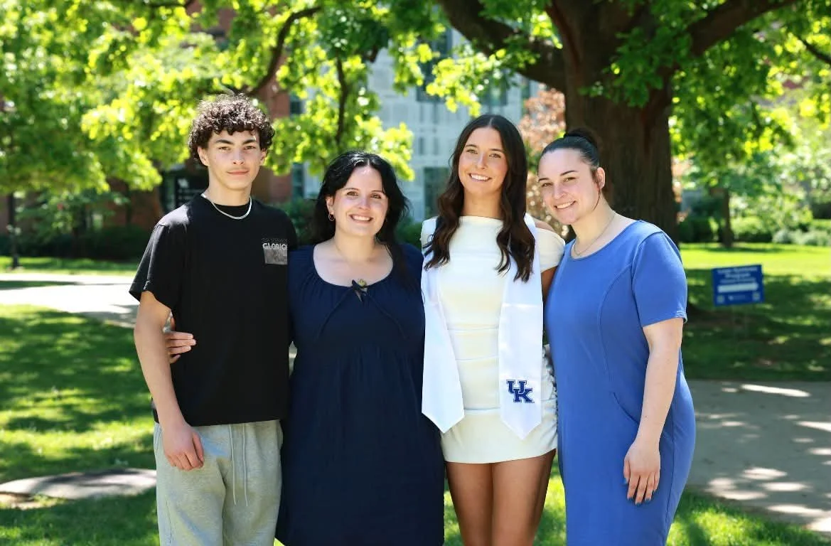 Four people, two women and two men, standing outdoors in a park on a sunny day, smiling at the camera. One woman is wearing a white graduation gown with a UK emblem. Behind them are green trees and a building in the background.