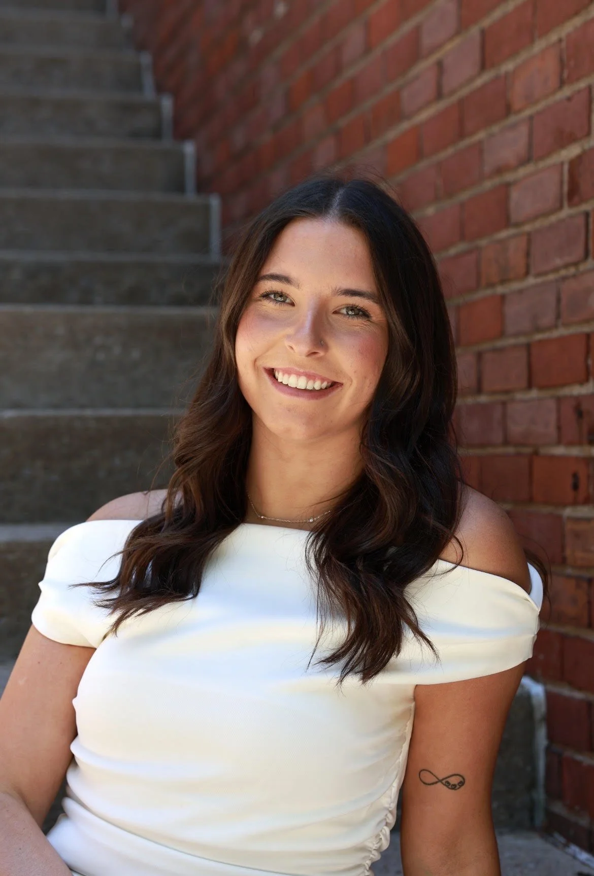 A woman with long dark hair, wearing a white off-the-shoulder top, smiling at camera, standing outdoors in front of a brick wall and concrete stairs.