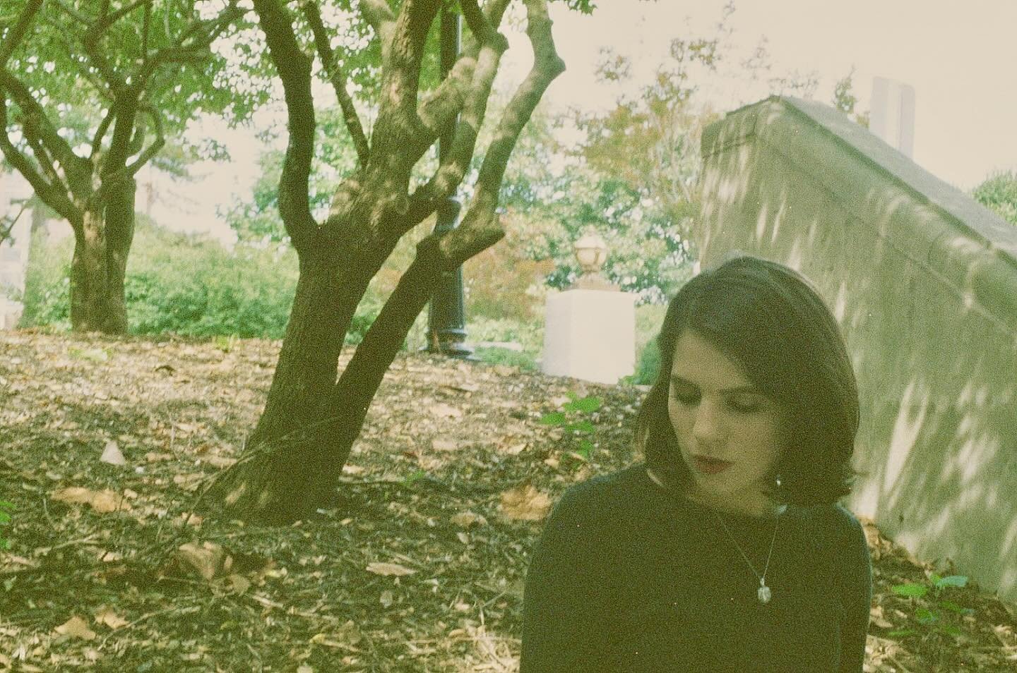 A woman with black hair and wearing a black top and necklace standing outdoors amidst trees and fallen leaves, with sunlight filtering through the branches.