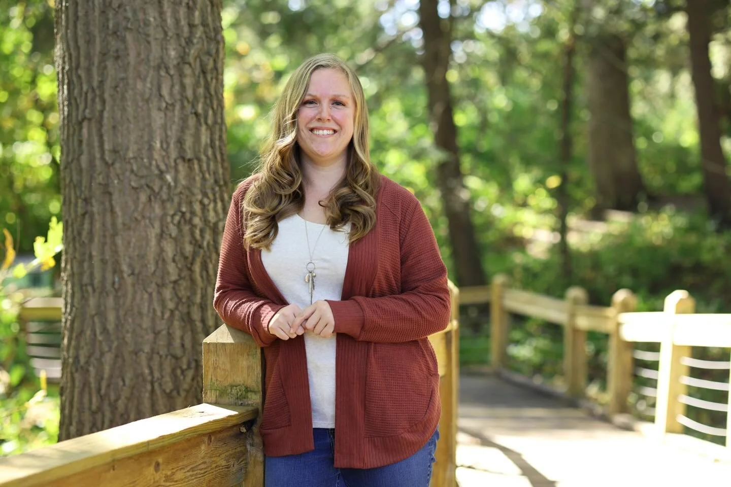 A young woman with long, wavy blonde hair standing on a wooden bridge in a forest, smiling at the camera.