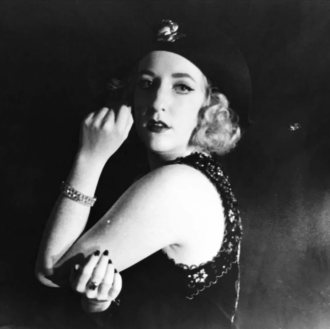 Black and white photograph of a woman with curly hair, wearing a wide-brimmed hat and an elegant dress with intricate details, posing thoughtfully with her hand near her face against a dark background.