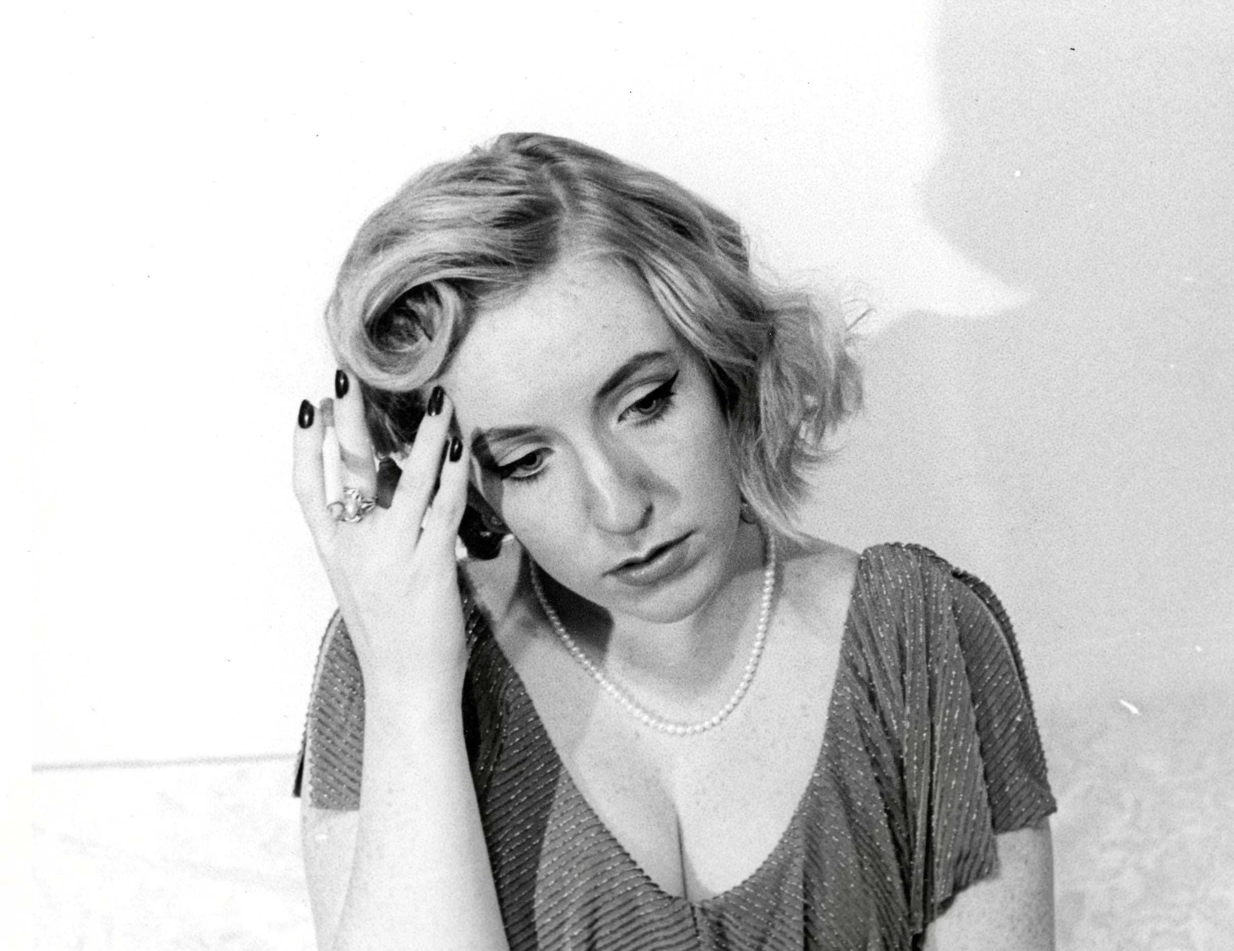 Black and white photo of a woman with vintage hairstyle, makeup, pearl necklace, and patterned dress, posing with her hand on her head.