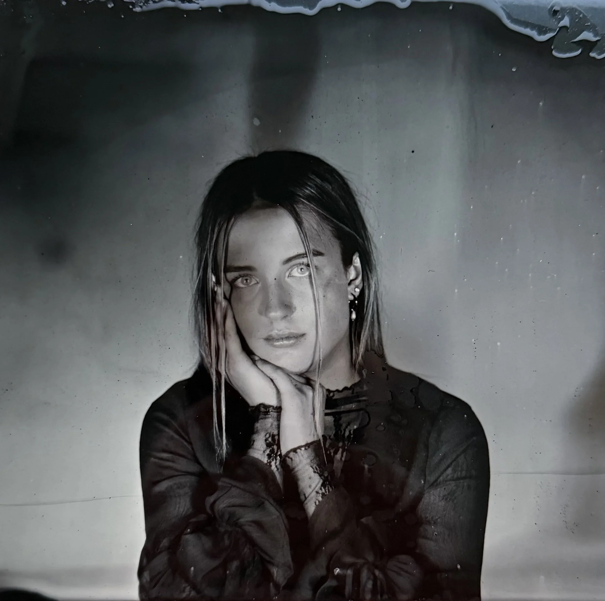 A person with long, wet dark hair and light skin laying down in water with their face partially visible, looking into the camera, with water dripping from their hair and some soap bubbles on the surface. The background is dark and the person appears to be in a bathtub or shower.