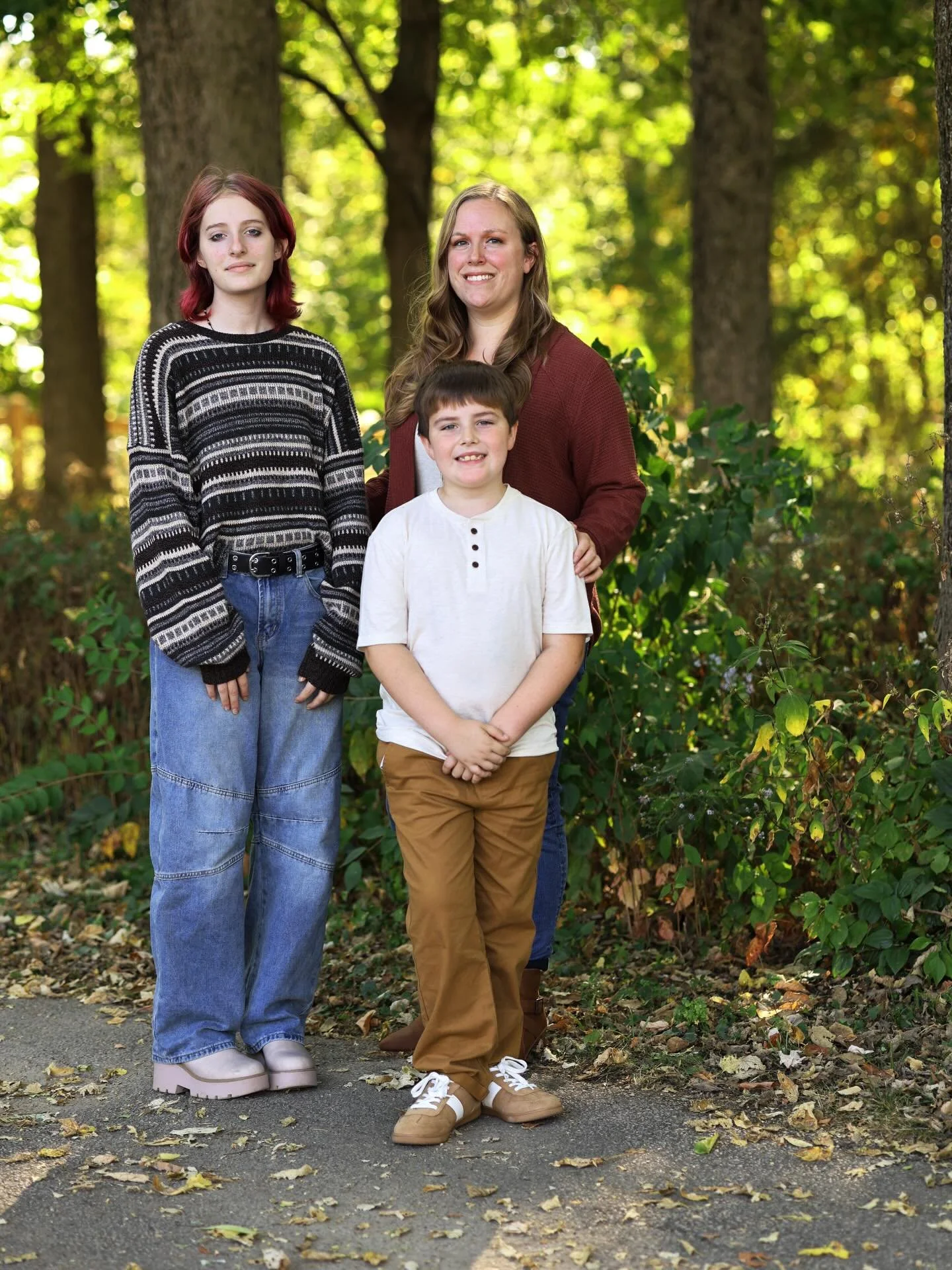 Three people, a teenage girl, a woman, and a young boy, standing outdoors on a sidewalk with trees and fallen leaves in the background.