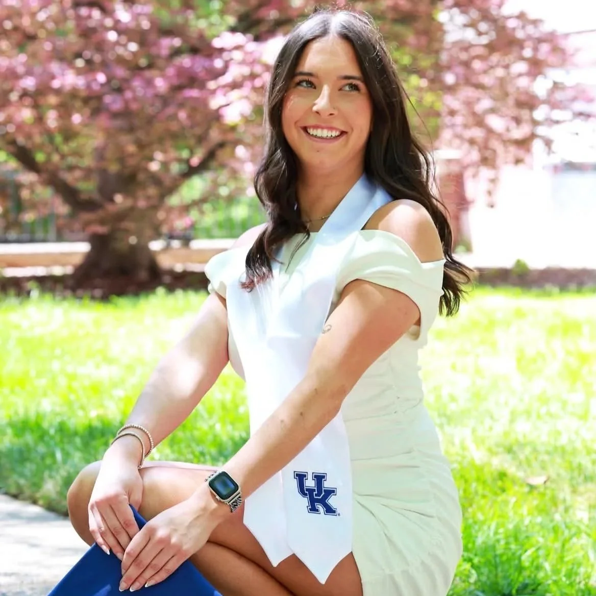 Young woman sitting outdoors on grass with pink blossoms in the background, smiling, wearing a white off-shoulder dress, a watch, and a University of Kentucky towel.