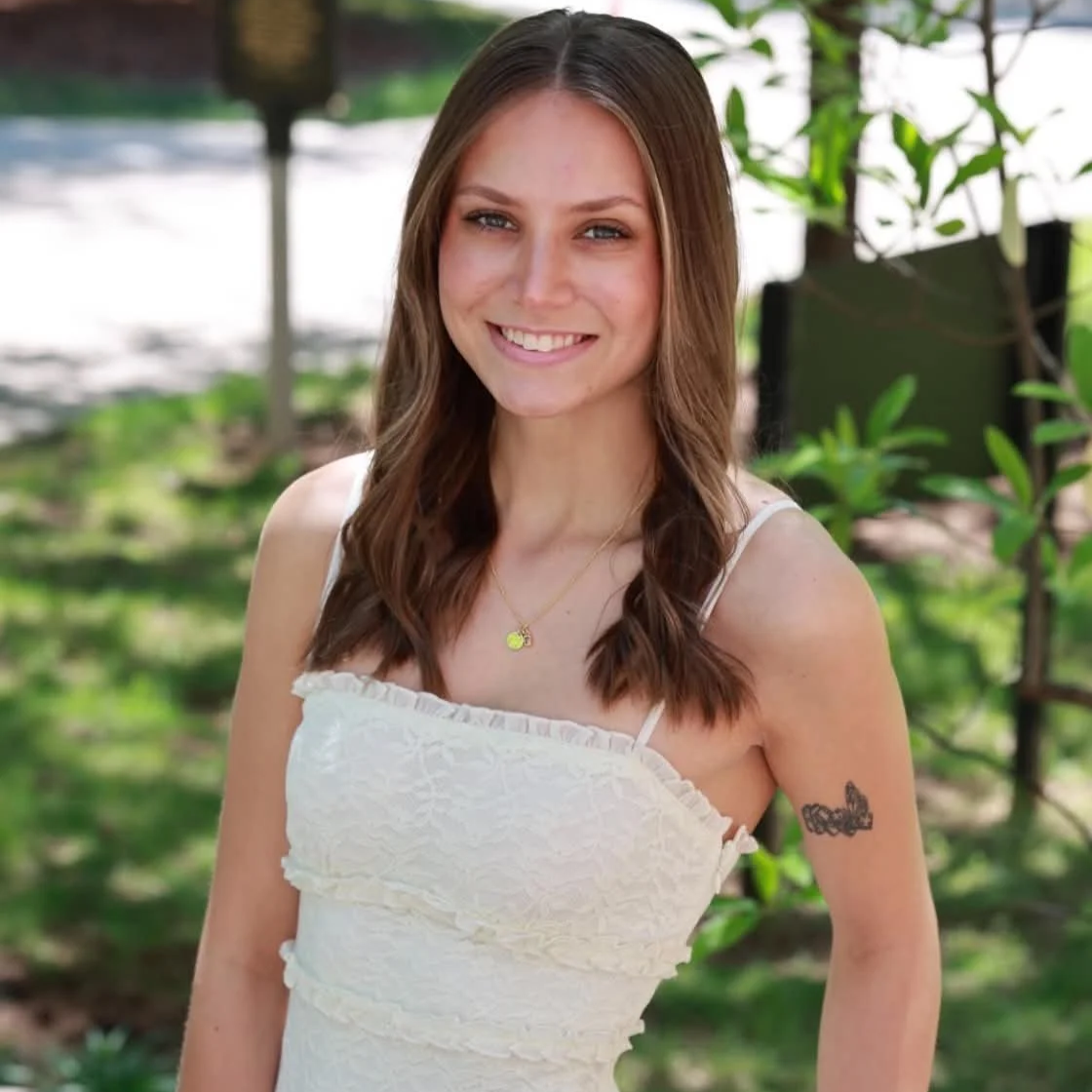 A young woman with long brown hair and blue eyes smiling outdoors, wearing a white sleeveless dress and a gold necklace with a round pendant, with greenery in the background.
