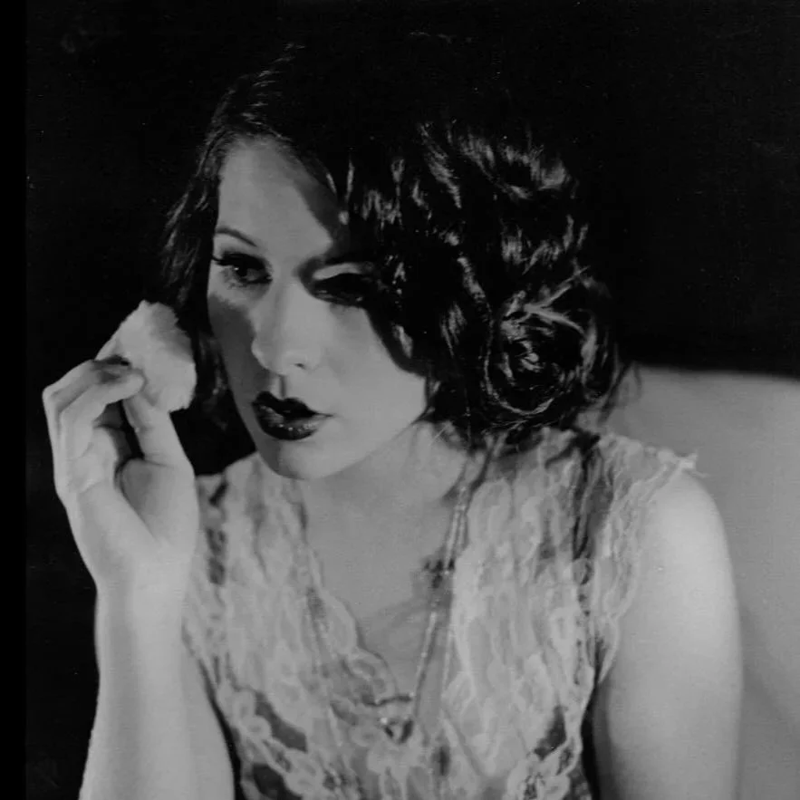 Black and white photo of a woman with short curly hair, wearing a lace top, holding a cloth to her face, with dark lipstick and eye makeup.