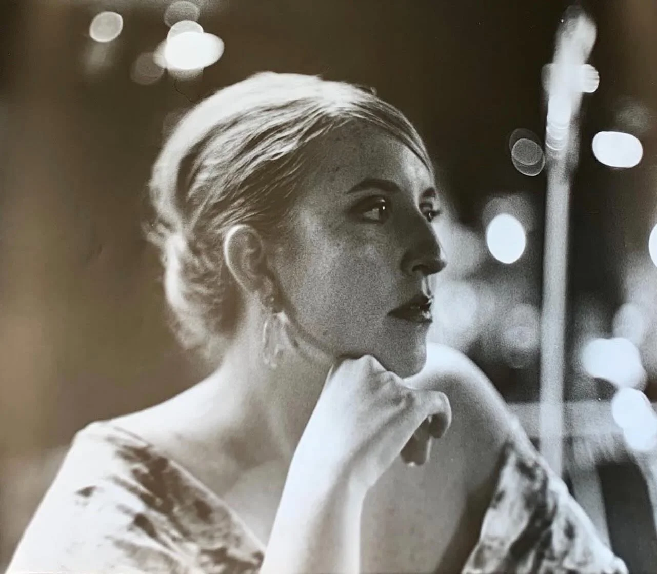 Black and white photograph of a woman with her hair styled in an updo, wearing earrings, gazing to the right with her hand resting on her chin, with blurred city lights in the background.