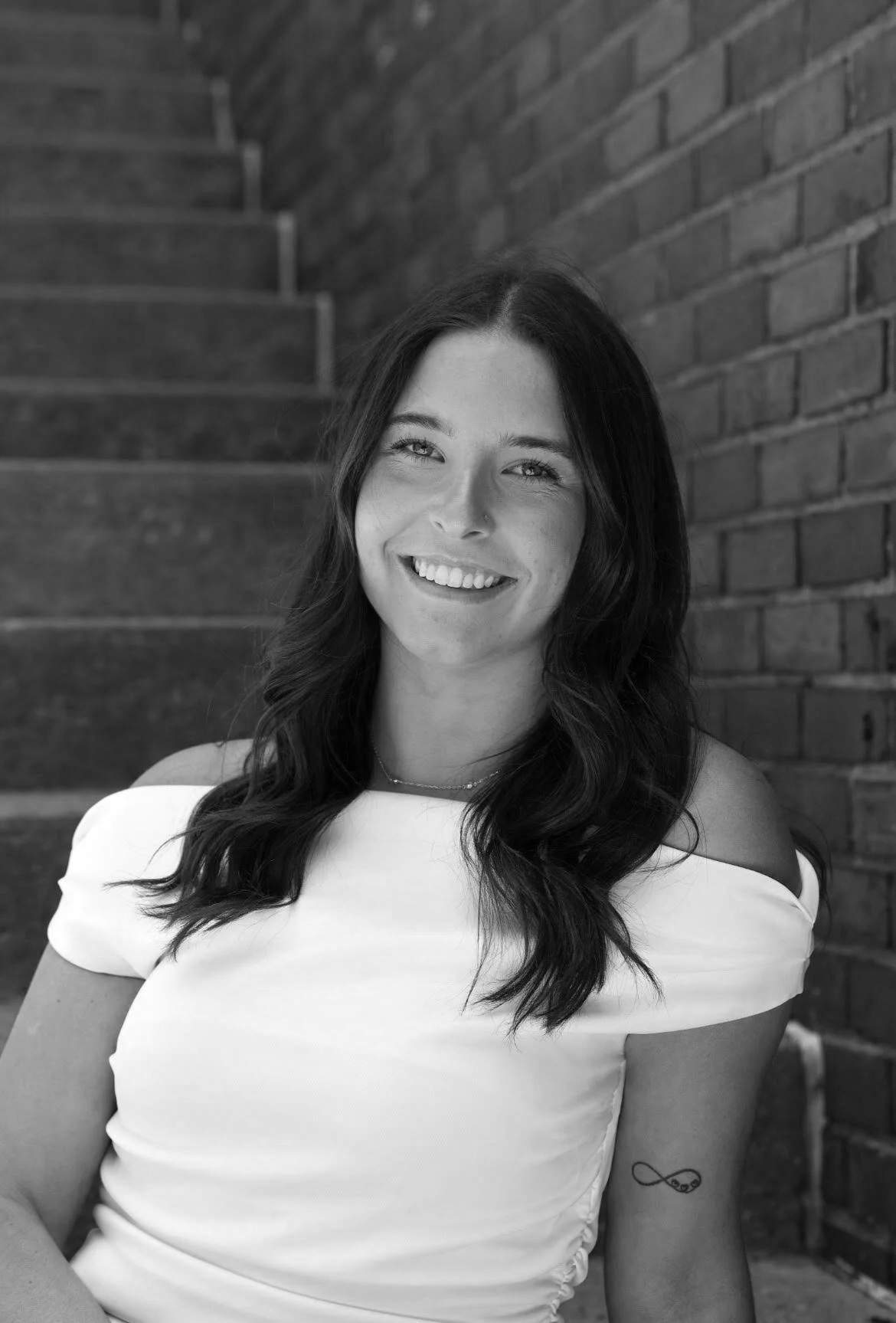 Black and white photo of a smiling woman with dark wavy hair, wearing an off-shoulder top, with a brick wall and staircase behind her.
