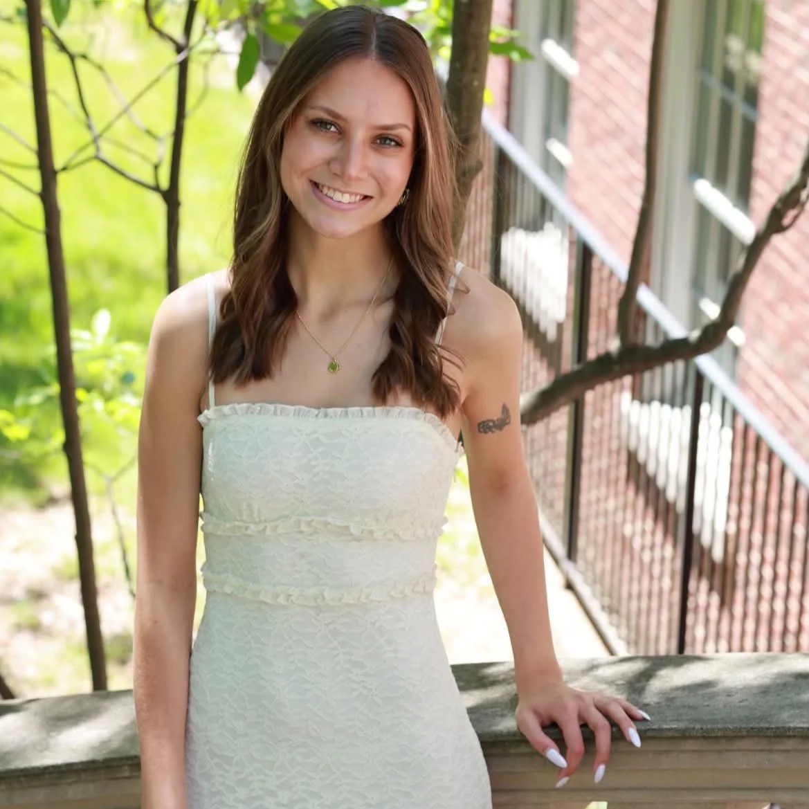 A young woman with brown hair and a tattoo on her arm, smiling outdoors on a balcony with greenery and a brick building in the background.