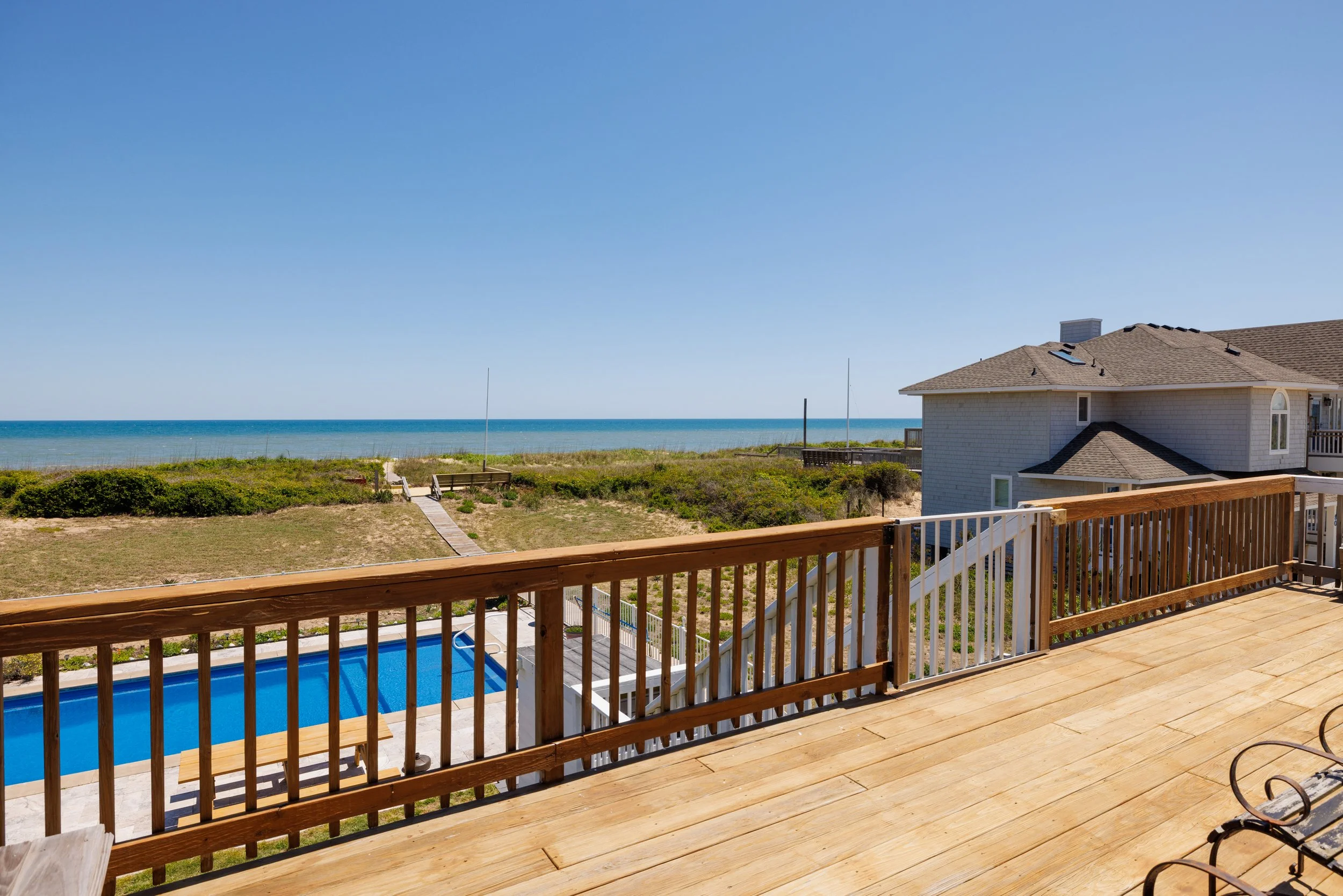Wooden deck with railing overlooking a backyard with a swimming pool, grassy area, and a house with a sloped roof near the beach and ocean in the background under a clear blue sky.