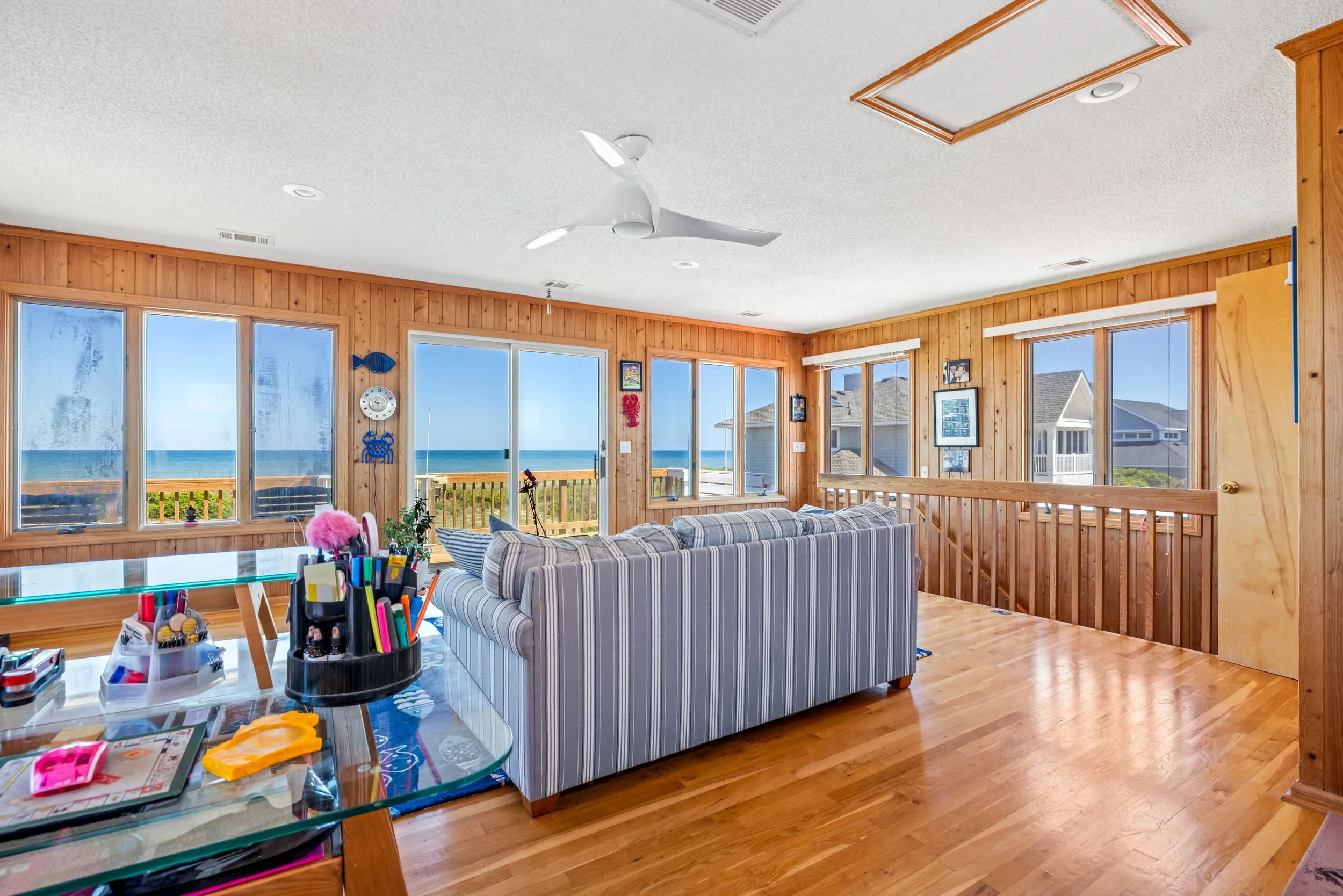 Living room with large windows overlooking the ocean, wooden walls, and a striped couch. There are various toys and art supplies on a glass table in the foreground, and decorative nautical items on the walls.