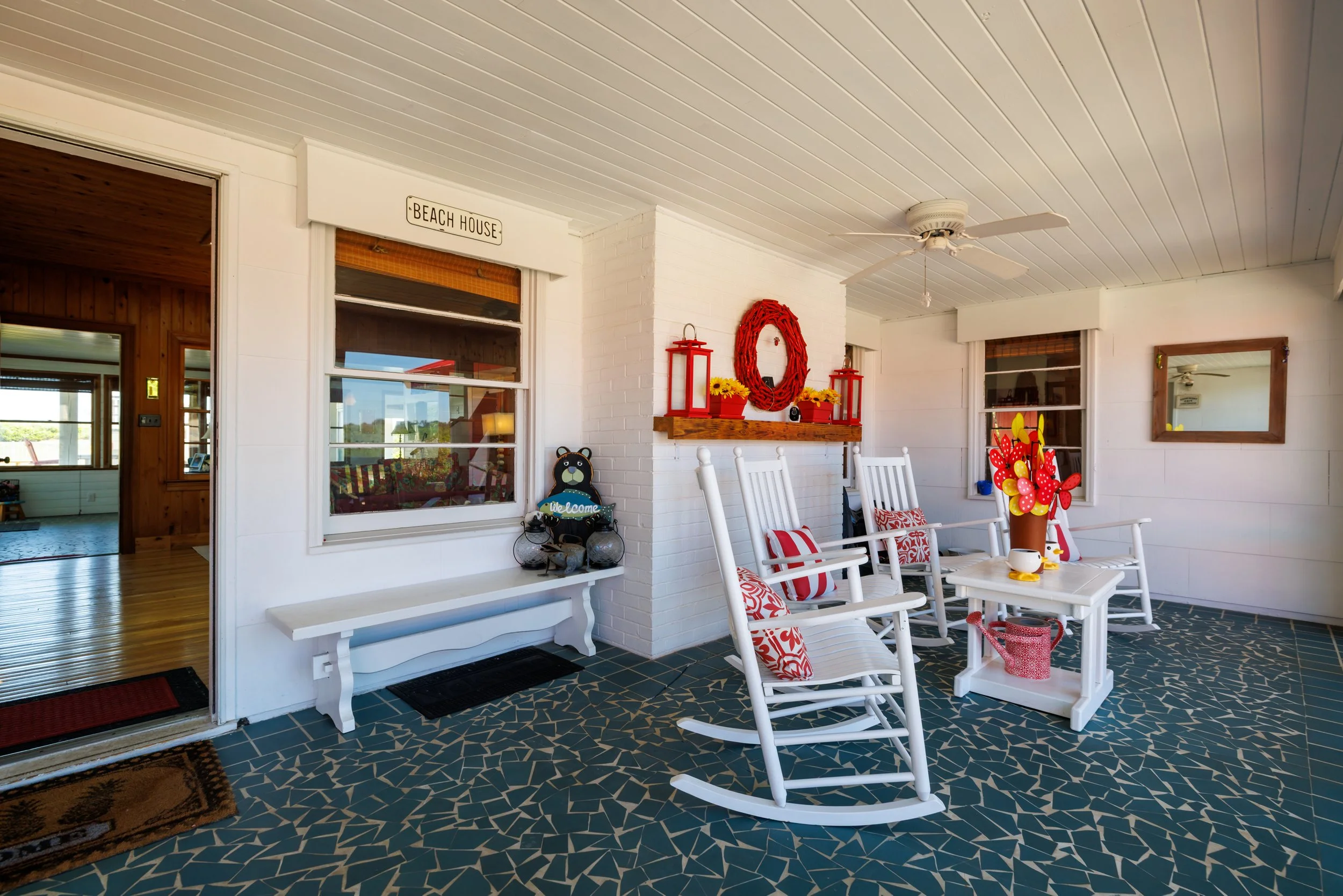 Porch area with white rocking chairs, a small white table, and colorful decorations including red lanterns, a wreath, and a Welcome bear figure. The floor has a dark tile pattern, and the walls are painted white with windows and doors leading inside.