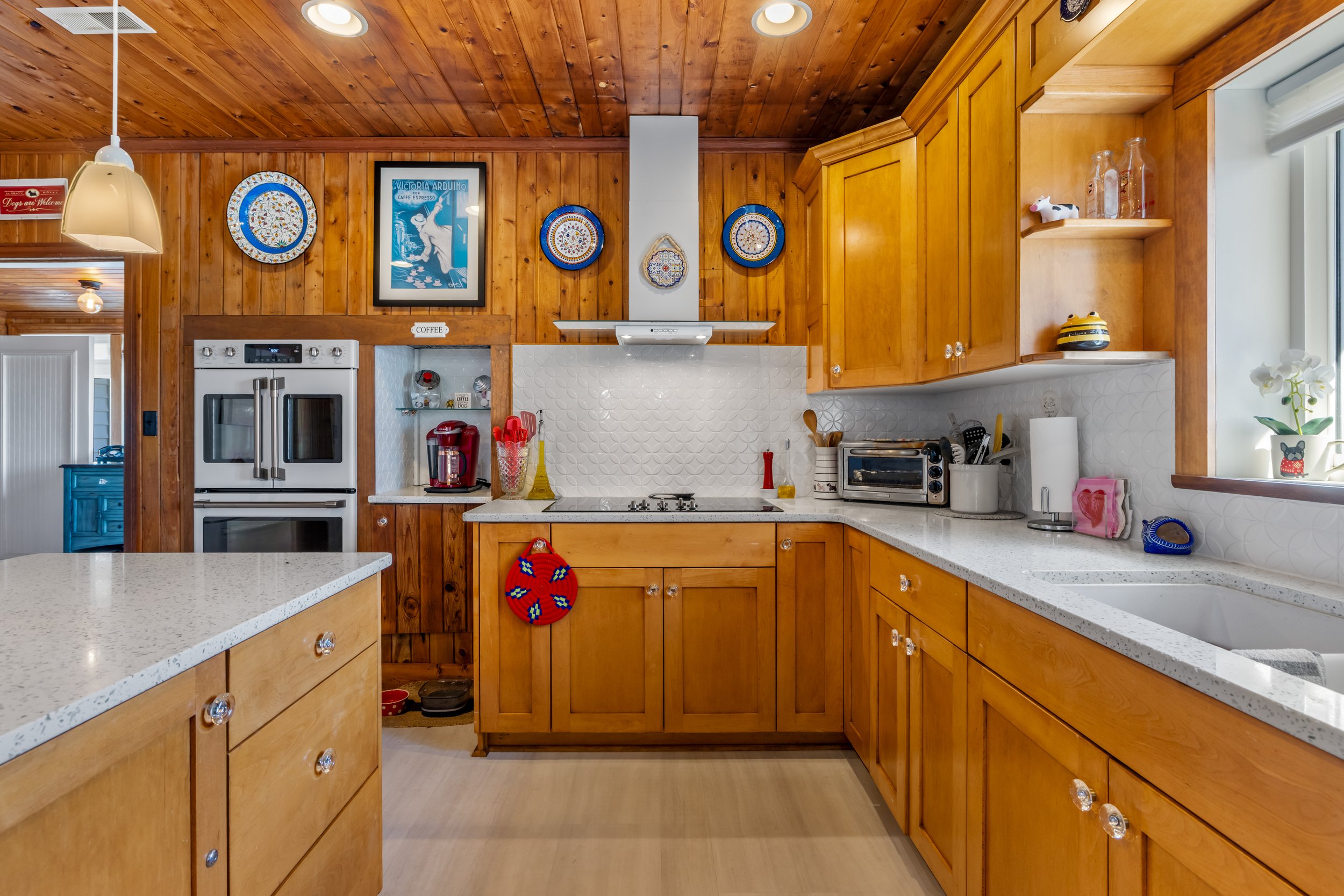A kitchen with wooden cabinets, a white countertop, and a wooden ceiling. Decor includes framed artwork and decorative plates on the wall, a red kitchen appliance, and various small items on the countertop.