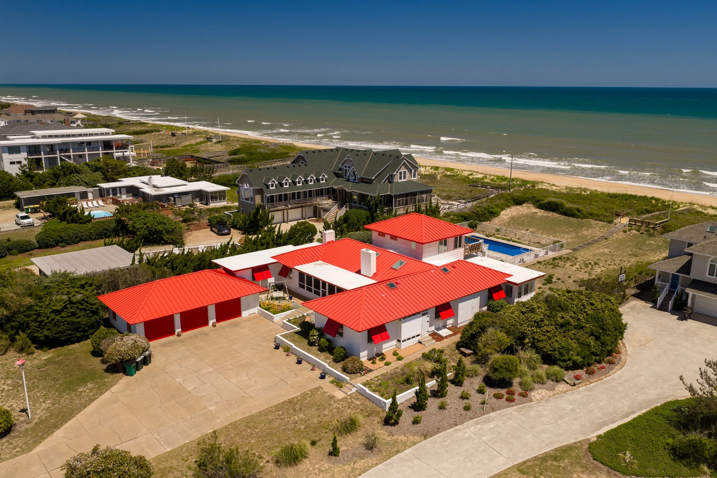 Aerial view of a coastal neighborhood with houses having red, gray, and white roofs, surrounded by greenery, with the beach and ocean in the background under a clear blue sky.