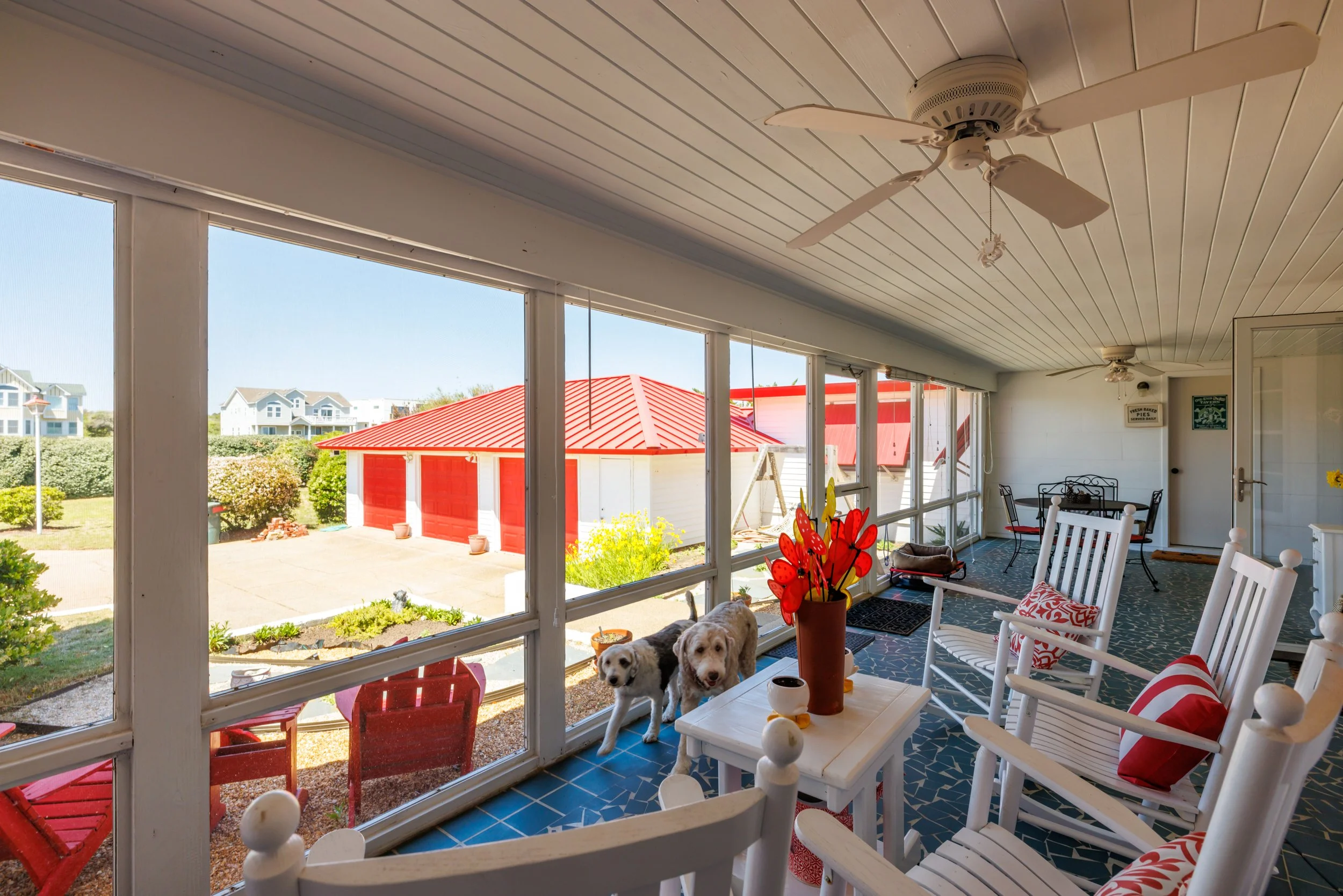 Covered porch with white wooden ceiling, ceiling fans, and large windows showing a bright outdoor yard with a red-roofed shed and houses in the distance. Inside, there are white chairs with red and white cushions, a table with decorative items, and t