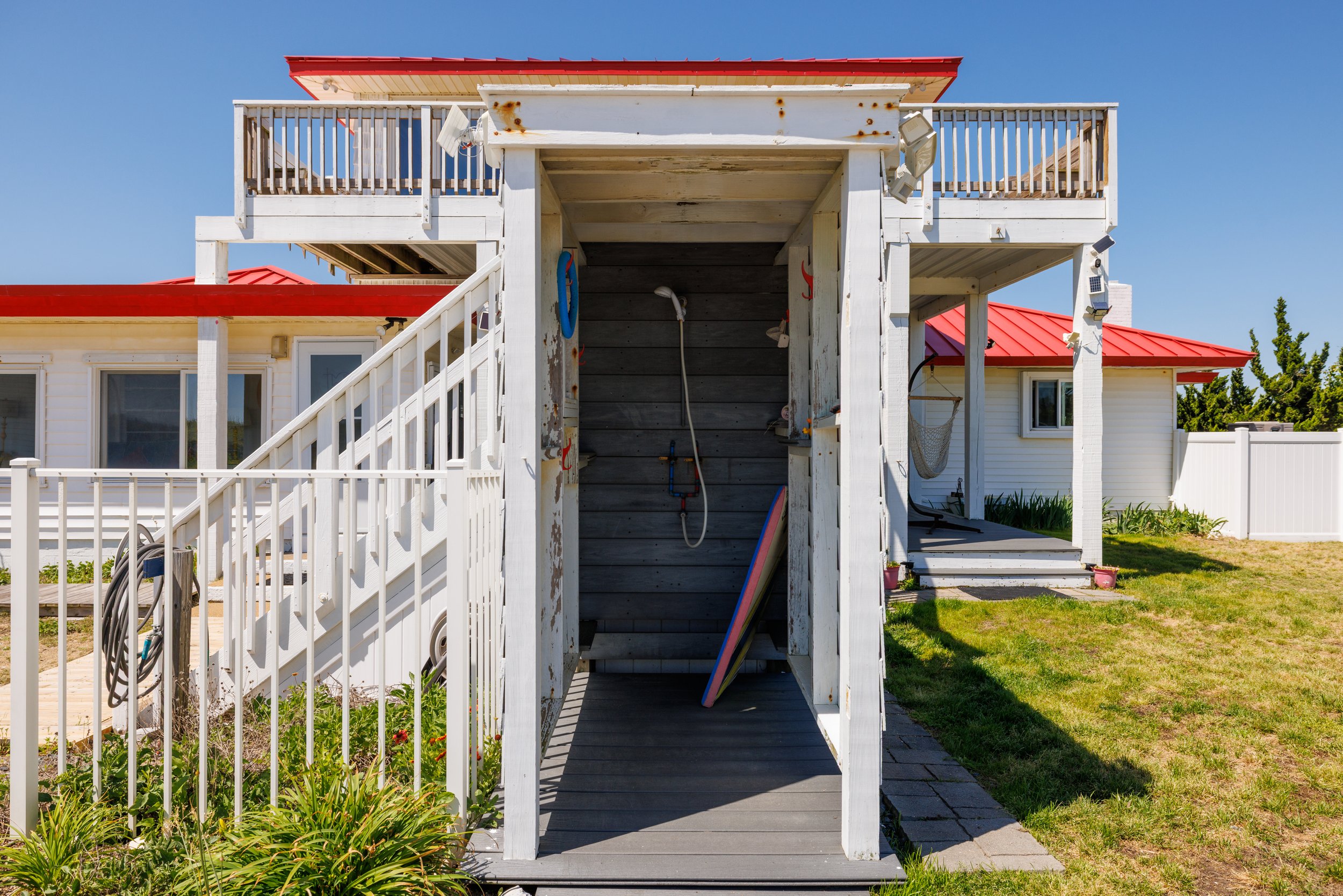Exterior view of a white wooden outdoor shower structure with a ladder, located in a backyard with a house featuring a red metal roof, white vinyl siding, and a white fence, surrounded by green grass and plants under a clear blue sky.