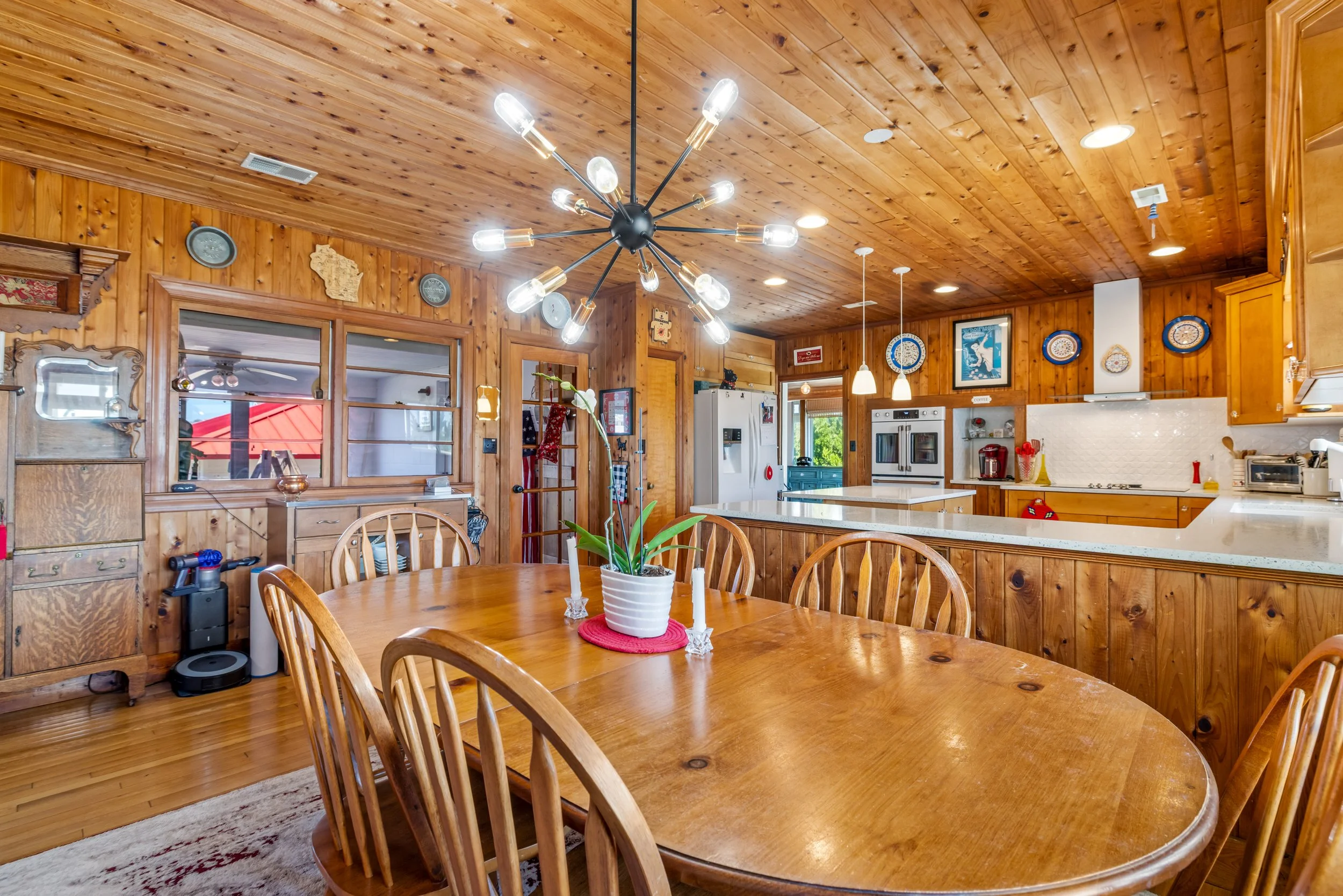 A wooden kitchen with a round dining table, centerpiece with orchids, and a modern black chandelier with exposed bulbs hanging from a wood-paneled ceiling.