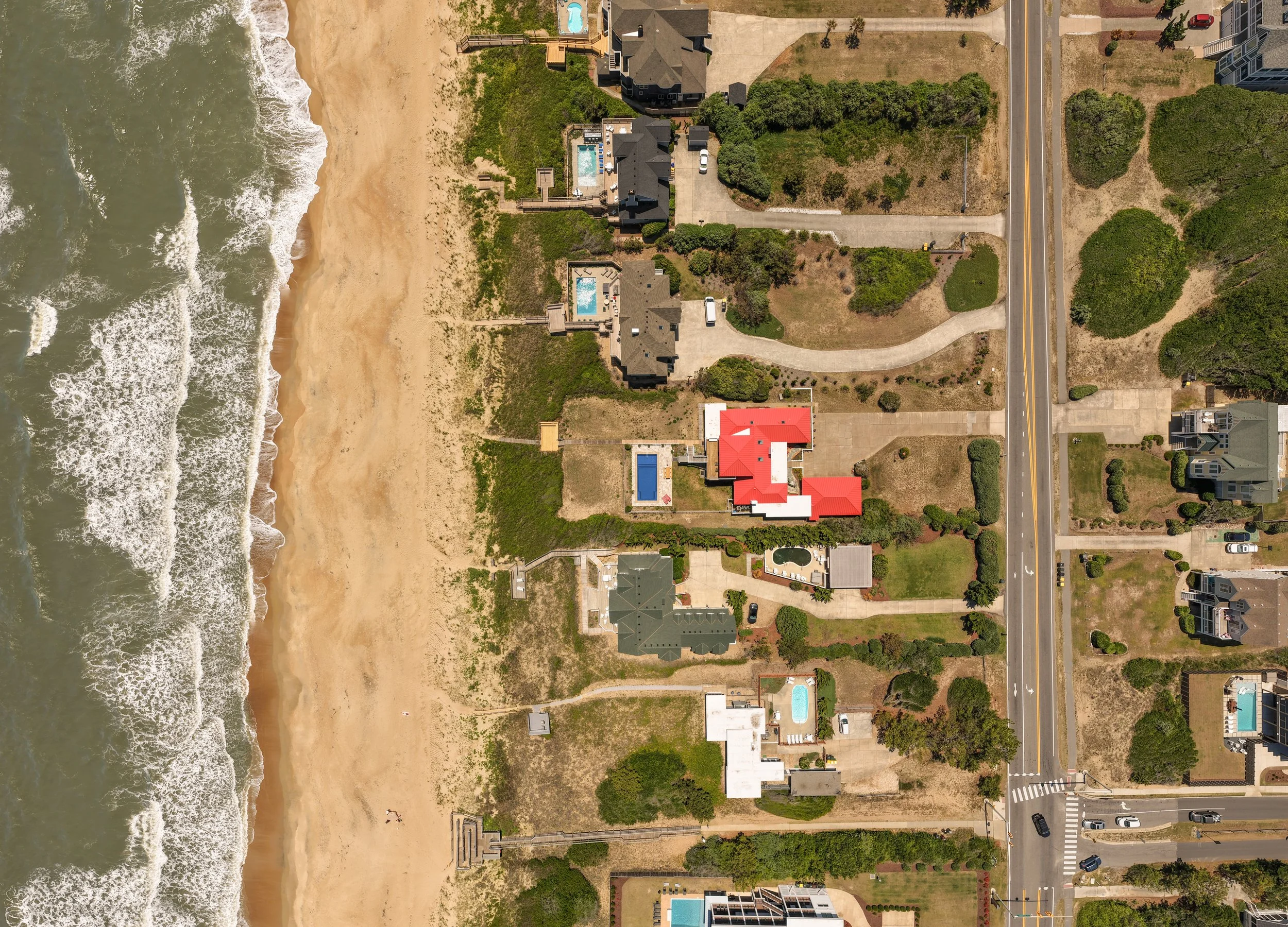 Aerial view of beachfront residential area with houses, swimming pools, greenery, and a sandy beach with ocean waves on the left side. A street runs vertically on the right side.