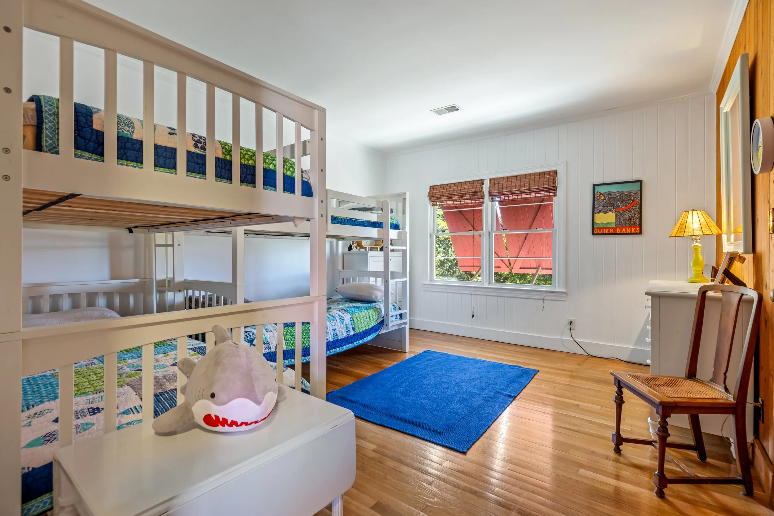 A bedroom with a bunk bed, a window with red blinds, a yellow lamp, a framed picture on the wall, and a plush toy shark on a white table.