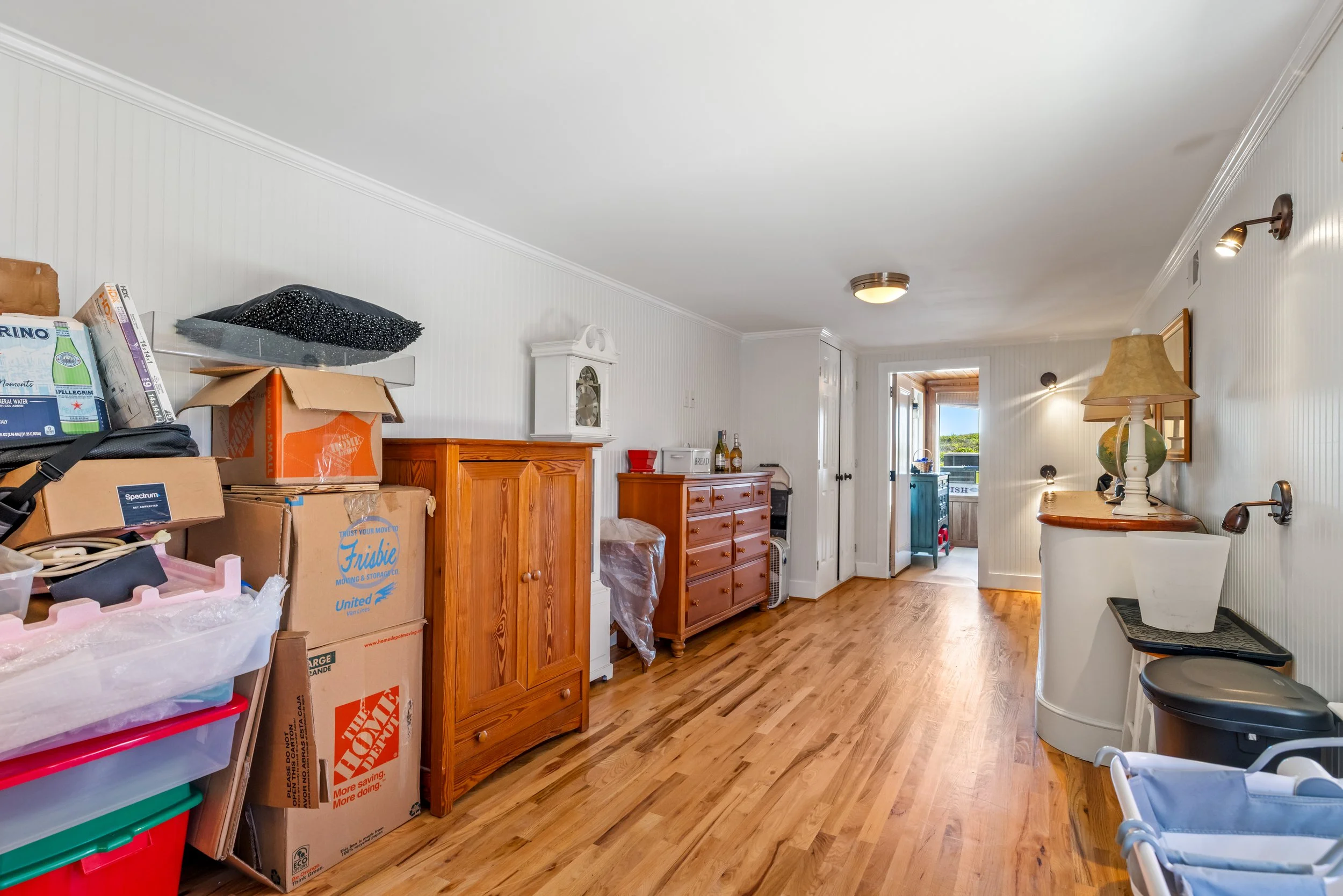 A cluttered living room with boxes, furniture, and household items, with a hardwood floor and white paneled walls.