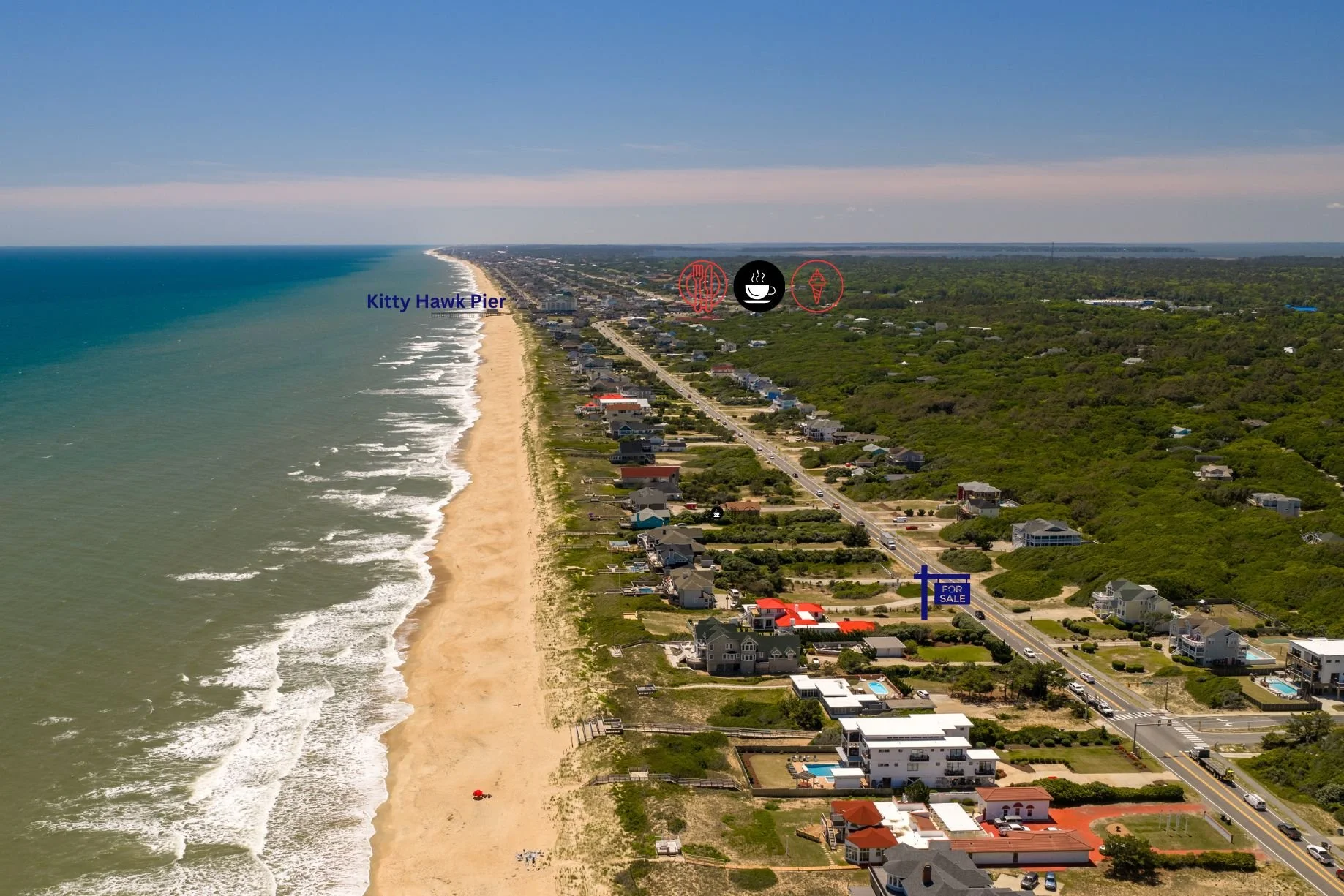 Aerial view of Kitty Hawk Pier along a sandy beach with ocean waves, beachfront houses, a road with traffic, and green vegetation, under a clear blue sky.