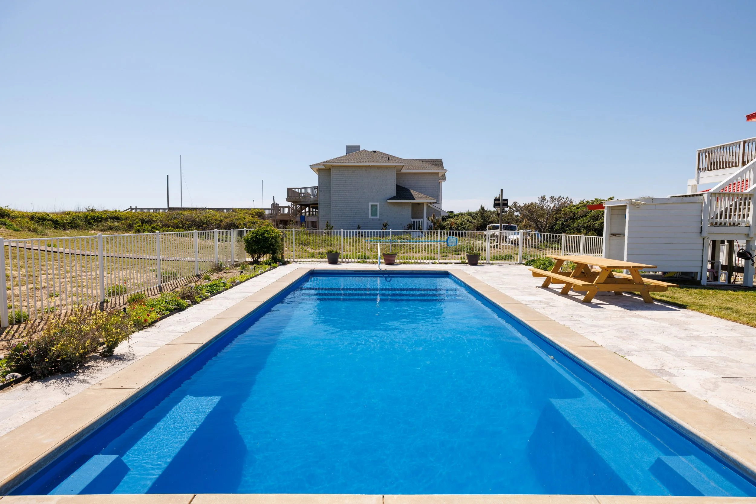 An outdoor backyard with a rectangular swimming pool, white fence, a picnic table, and a two-story house under a clear blue sky.