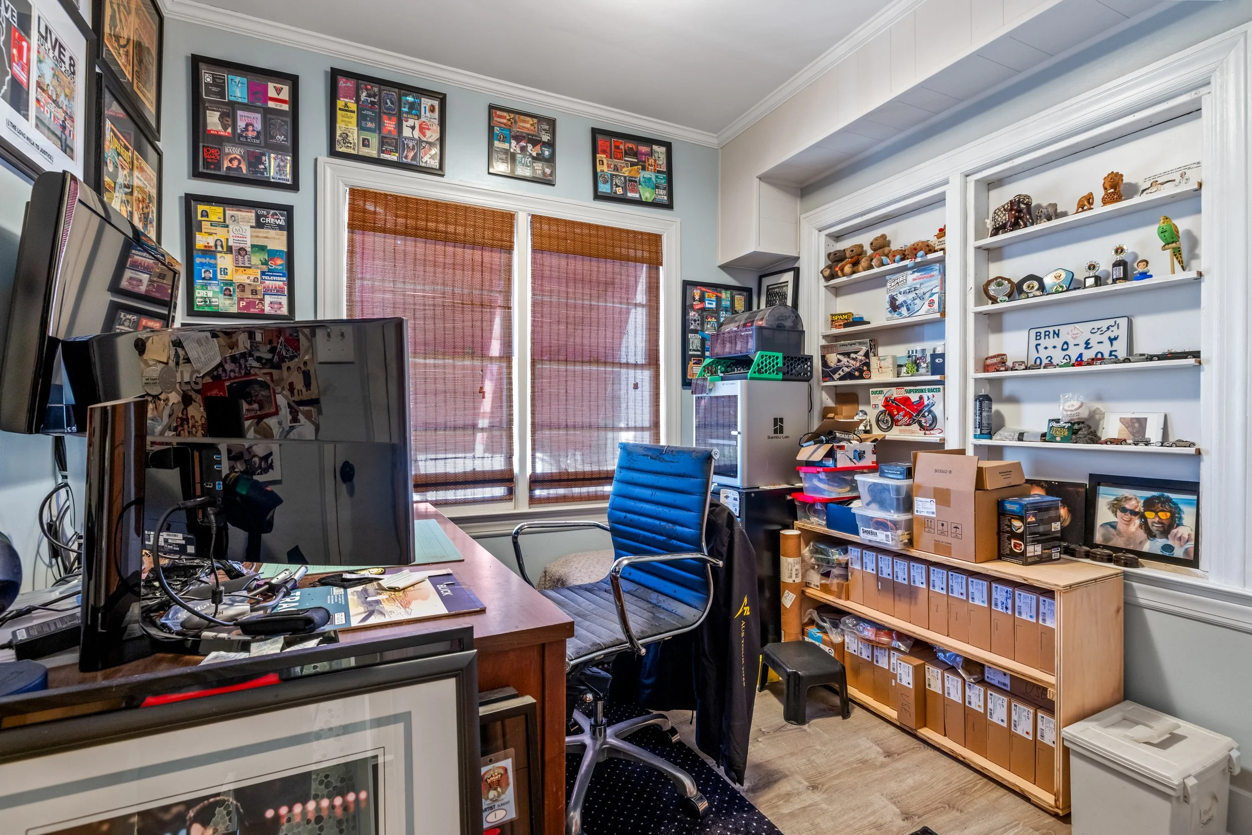 A cluttered home office with a wooden desk containing computer monitors, wires, and papers. Behind the desk, a blue office chair is positioned. The wall has a collage of framed pictures and certificates. There are large windows with brown blinds, and