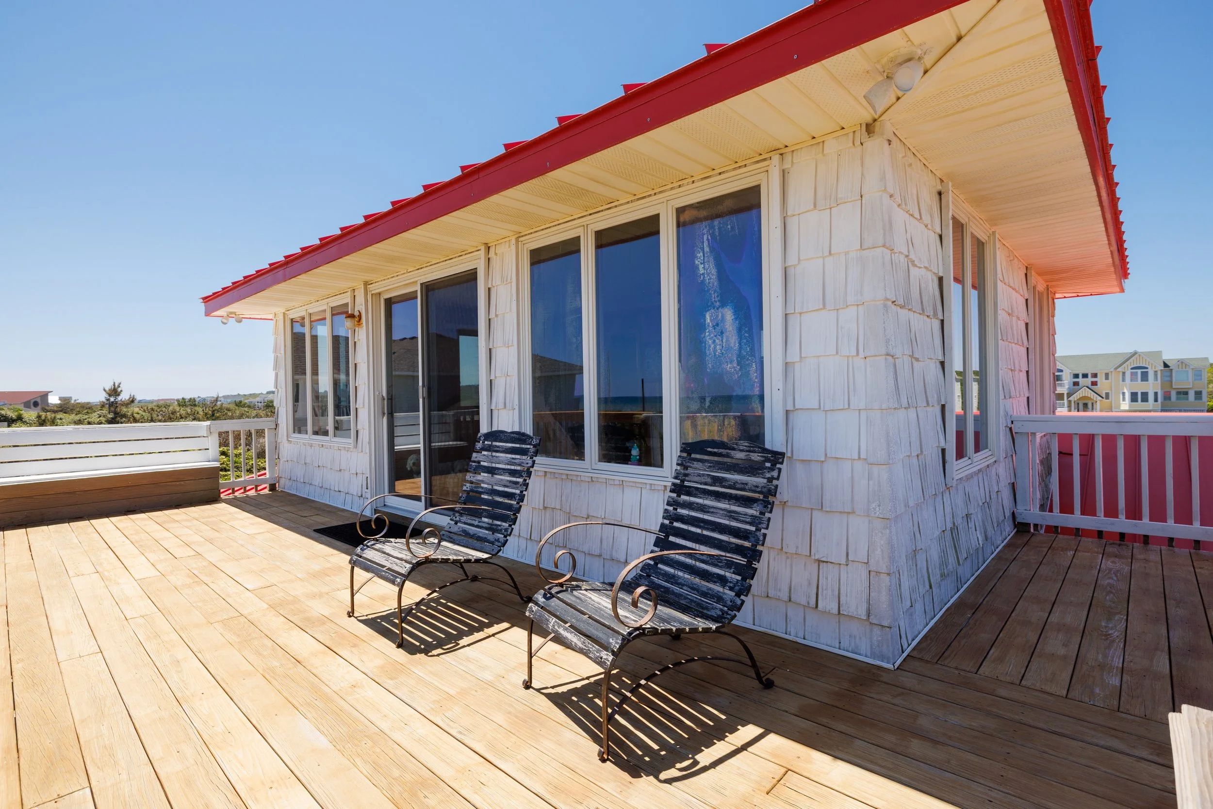 Balcony or terrace with wooden flooring, two black metal chairs, and a building with beige shingle exterior and large windows. In the background, there are other buildings and a clear blue sky.