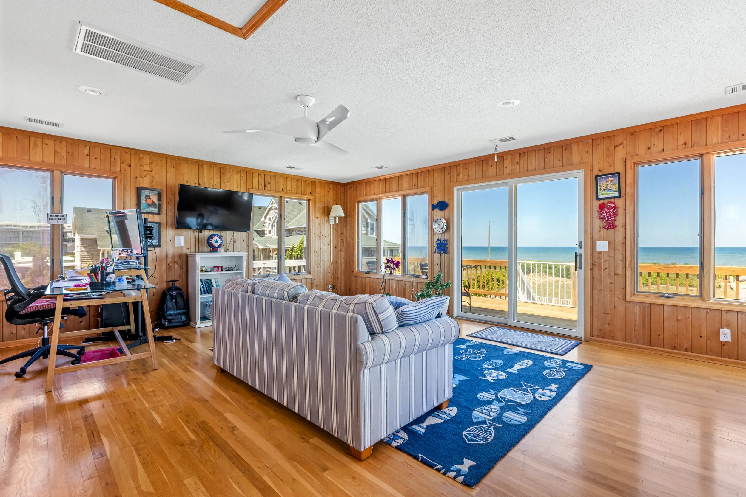 Living room with wooden walls and large windows showing a beach view, striped sofa, flat-screen TV, ceiling fan, and a blue area rug with shells pattern.