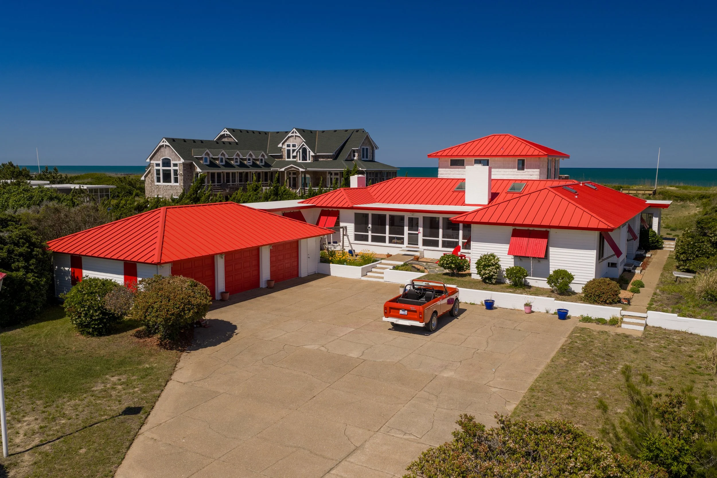 View of a coastal residential area with two houses featuring red roofs, a driveway, greenery, and a background of the ocean and clear blue sky.