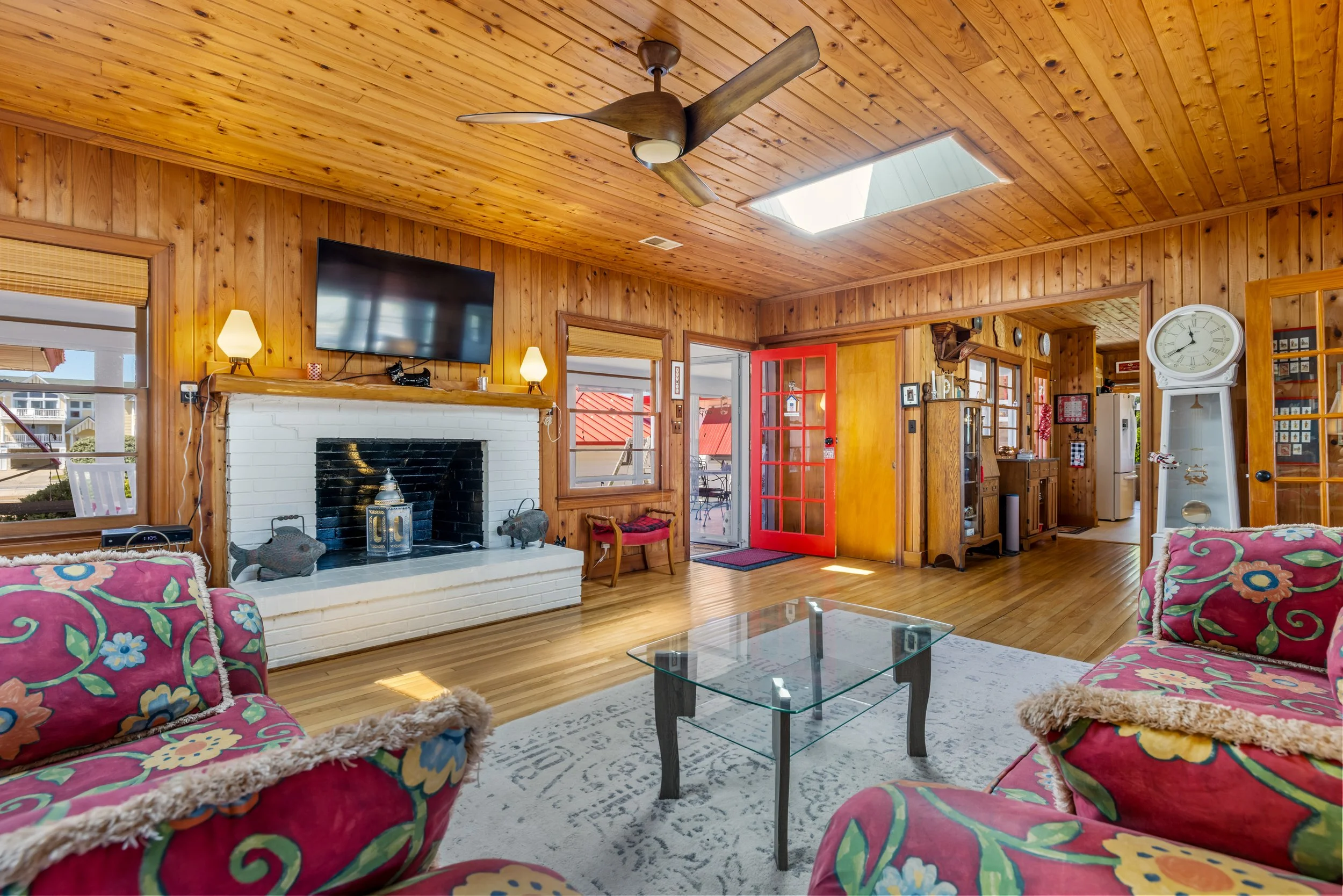 A cozy living room with wood-paneled ceiling and walls, a white brick fireplace, a mounted flat-screen TV, a ceiling fan, a glass coffee table, and colorful armchairs with floral pillows. There is a large window with blinds and a skylight.