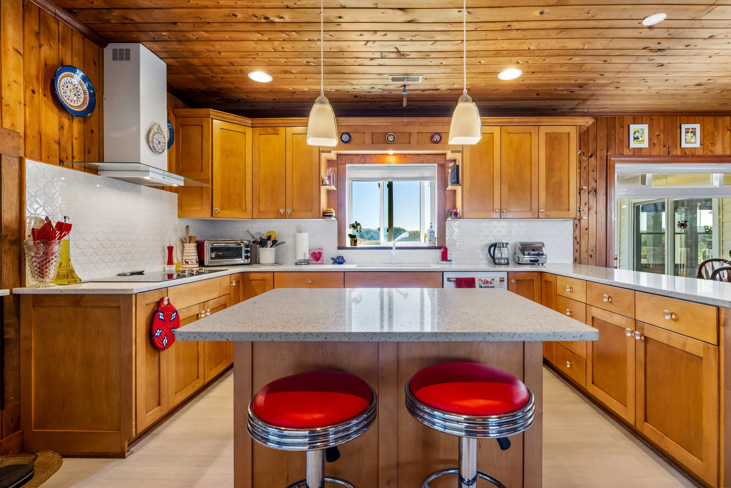 A kitchen with wooden cabinets and walls, a white countertop, and a window above the sink. The kitchen has hanging pendant lights and various small decorative plates on the wall. There are red and black barstools at an island in the foreground.