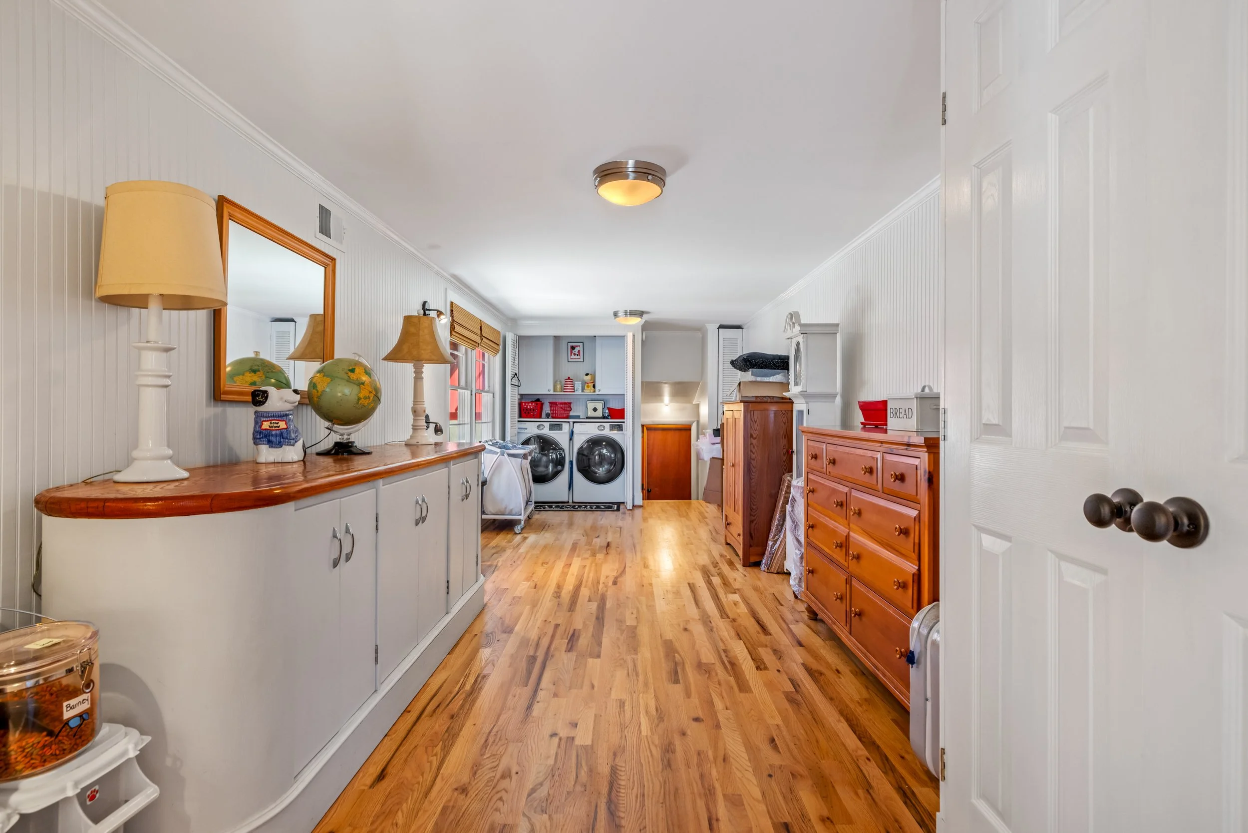 A laundry room with white beadboard walls, wooden floors, a white cabinet with decorative lamps and a globe on top, a washer and dryer set, wooden storage cabinets, and shelves with baskets and laundry supplies.