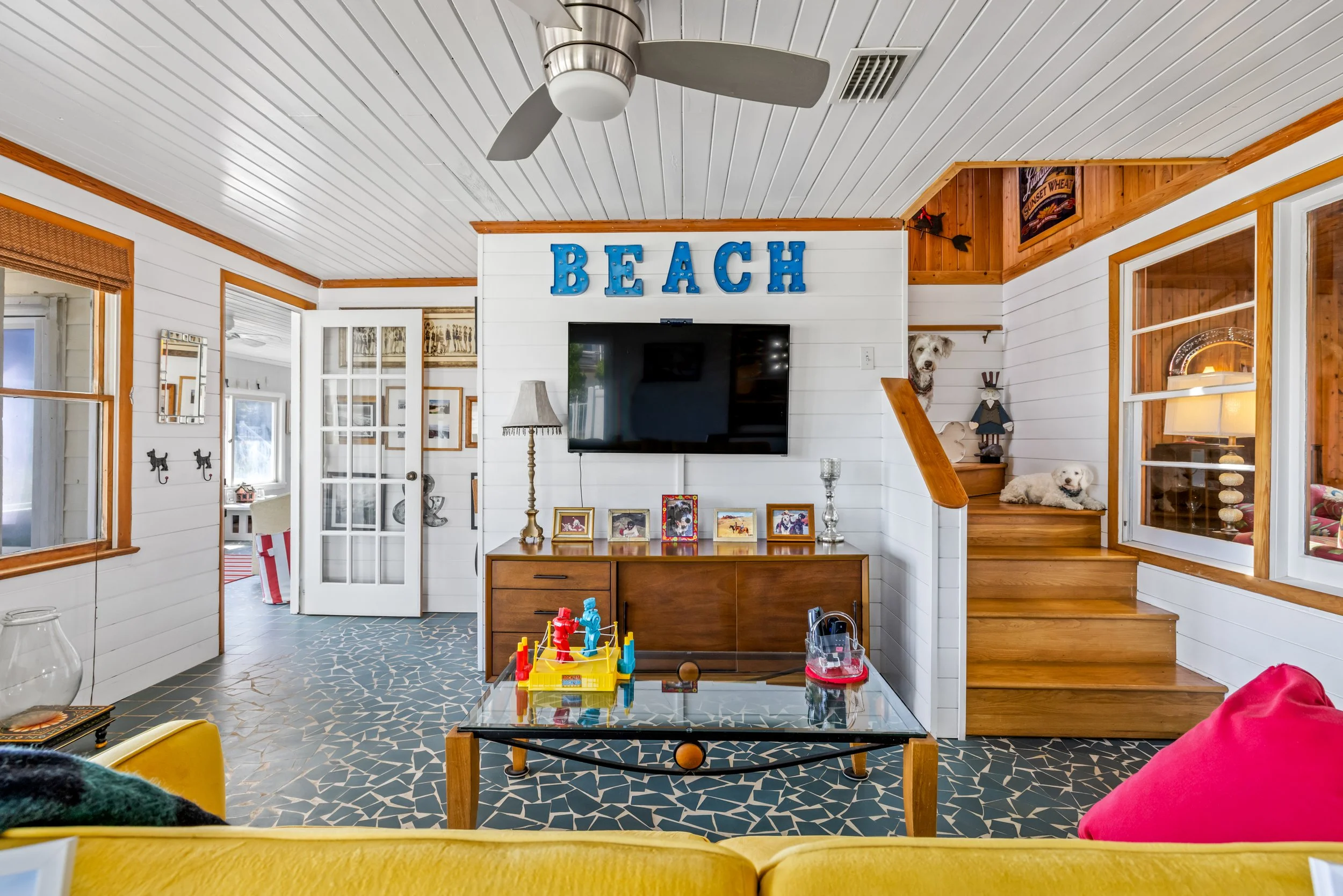 Inside a beach-themed living room with white wooden walls and ceiling, featuring a ceiling fan, a wall-mounted television, framed photos on a wooden cabinet, and two white dogs on a wooden staircase.